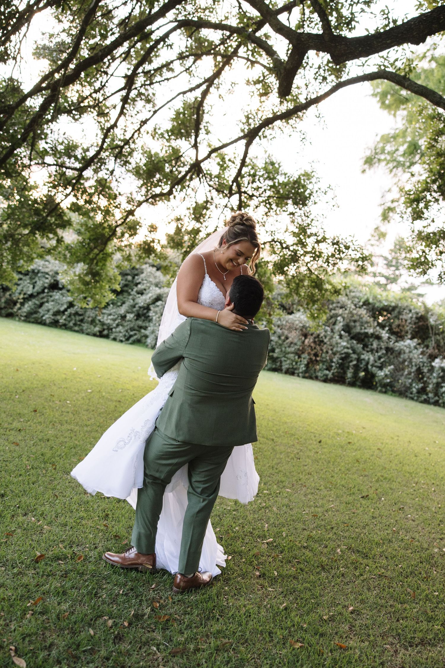 Groom in green suit lifts bride in white wedding dress outdoors under oak tree on lush green lawn.