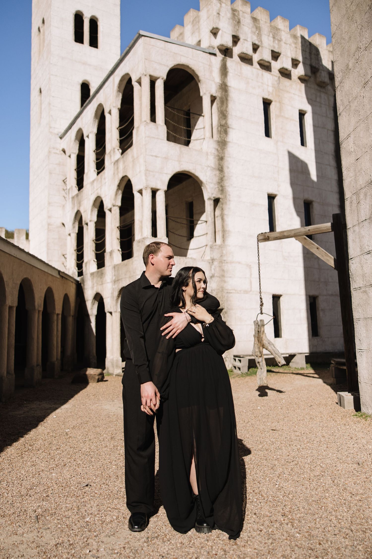 Couple dressed in black standing together before a historic white stone castle in sunlight.