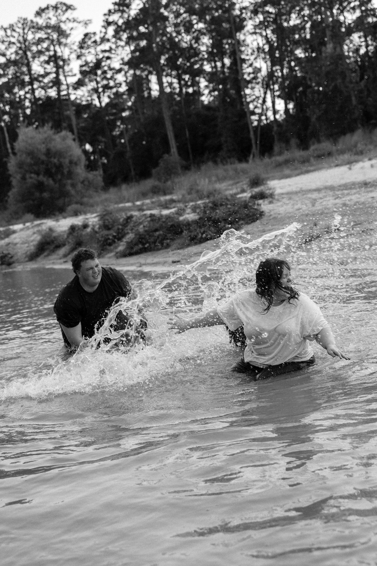 Black and white photo of two people splashing and playing in a river with trees in the background.