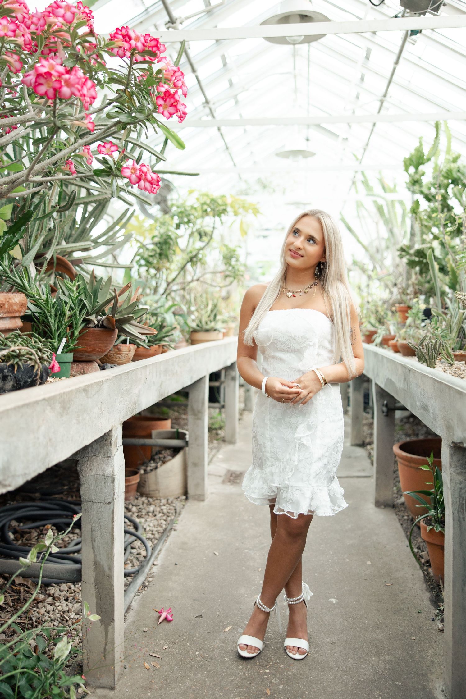 Individual in white dress poses among pink flowers in a greenhouse setting.