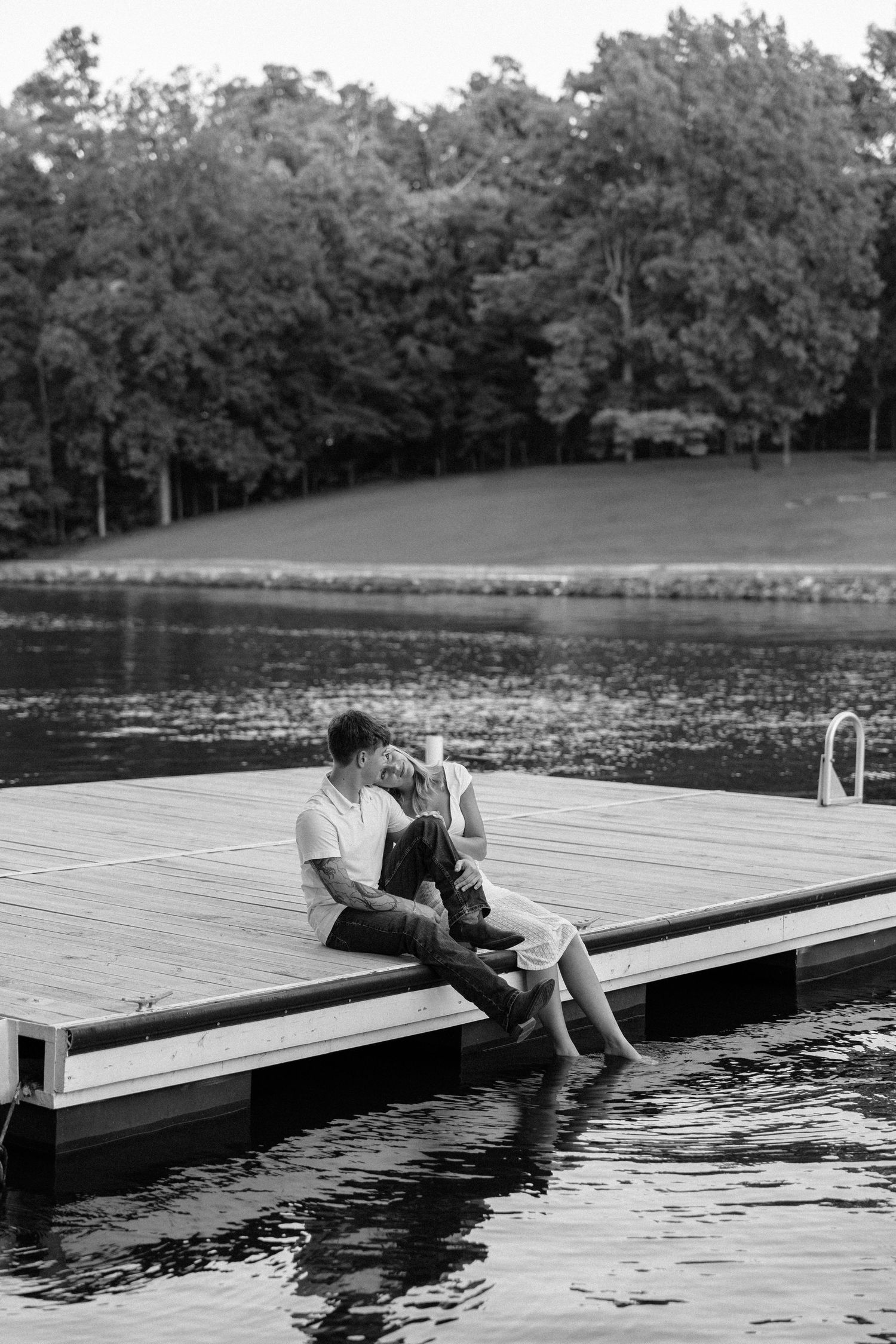 Black and white photo of a couple sitting closely on a wooden dock by a lake, surrounded by lush trees.