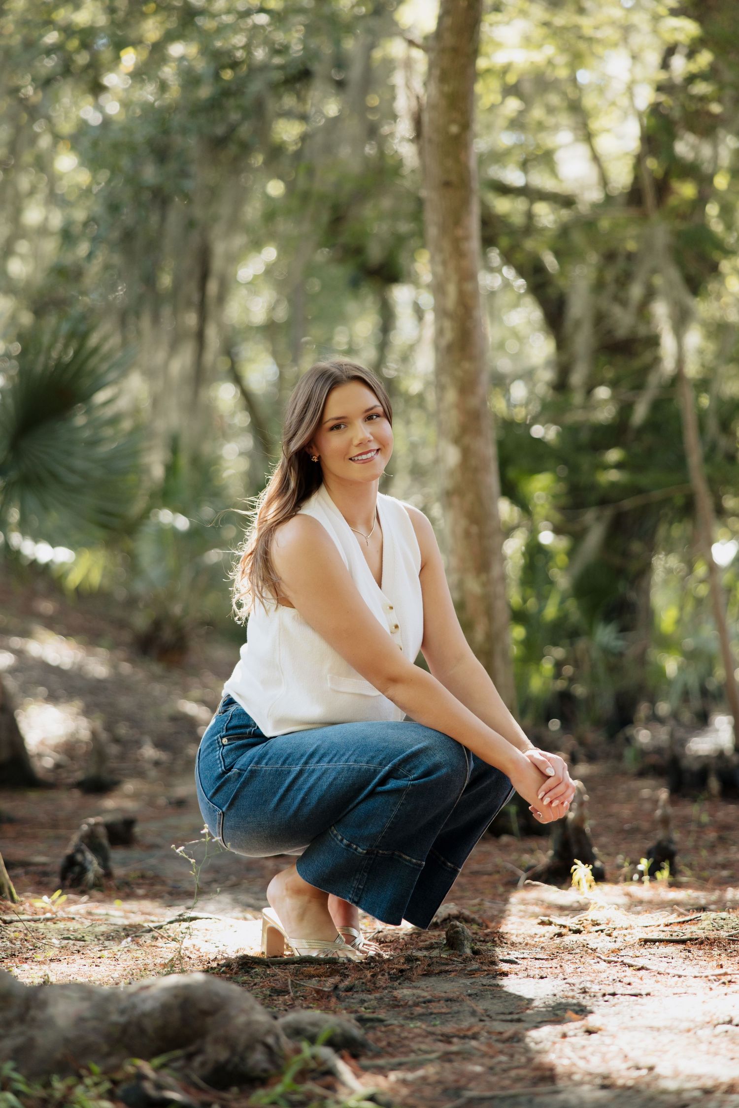 A person in a white top and jeans crouches in a sunlit forest setting with dappled light filtering through trees.