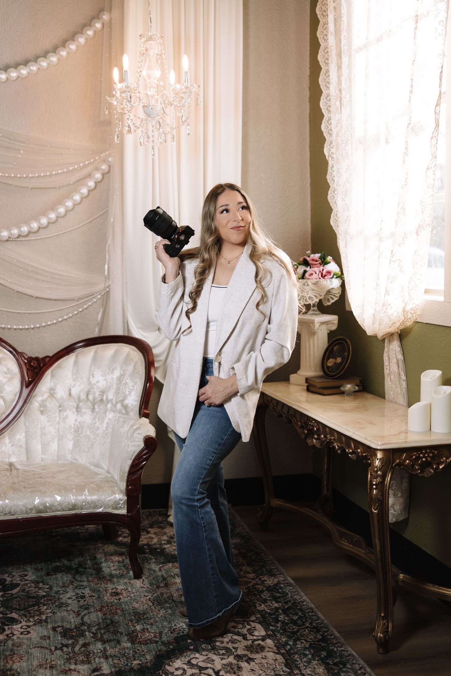 A photographer in a white blazer and jeans holds a camera in an elegant room with vintage furniture and chandelier.