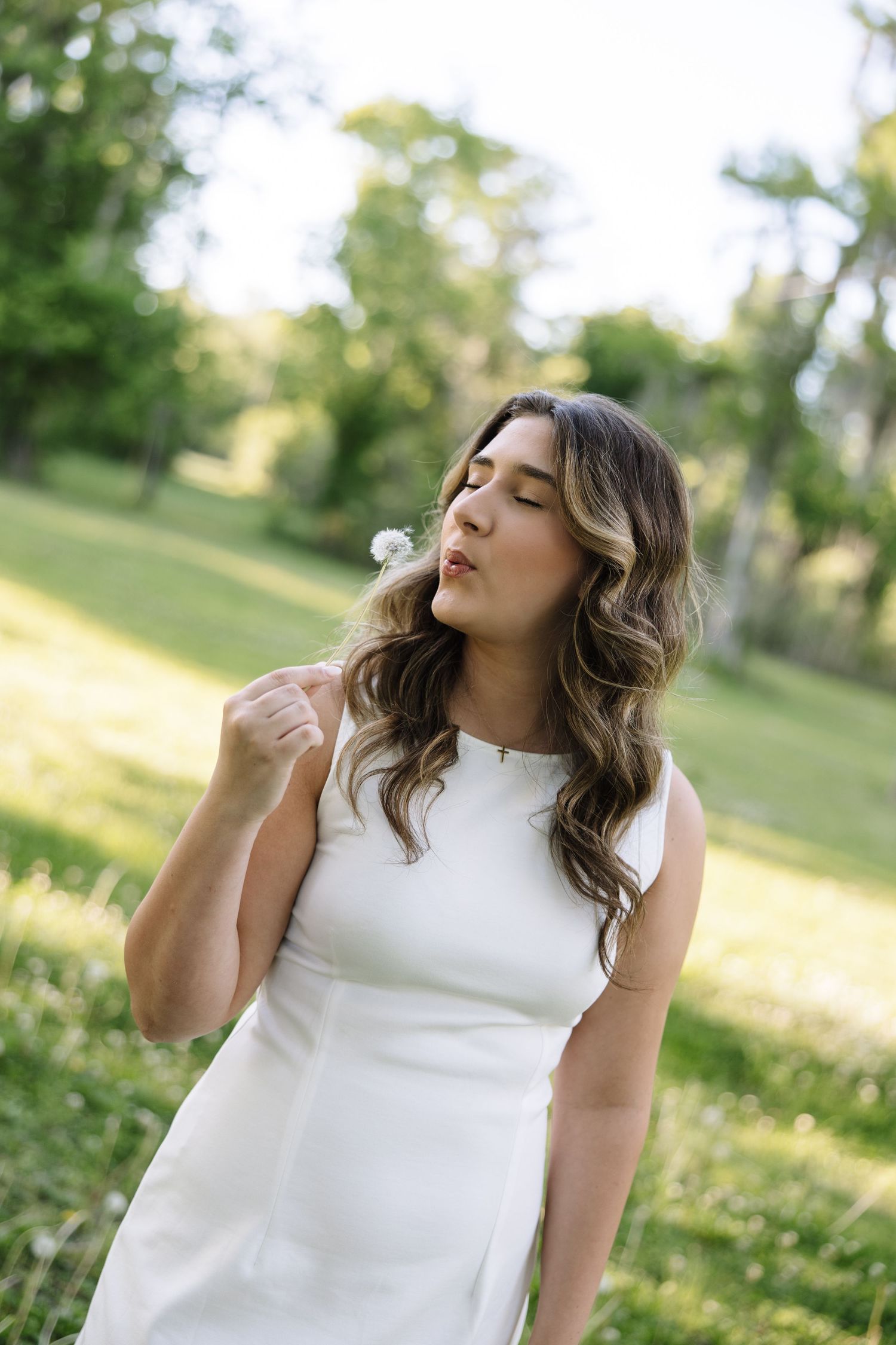 Woman in white sleeveless dress blowing a dandelion in a lush green park on a sunny day.