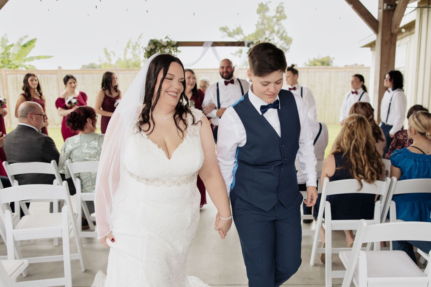 A joyful couple walks down the aisle together at an outdoor wedding ceremony with guests seated in white chairs.