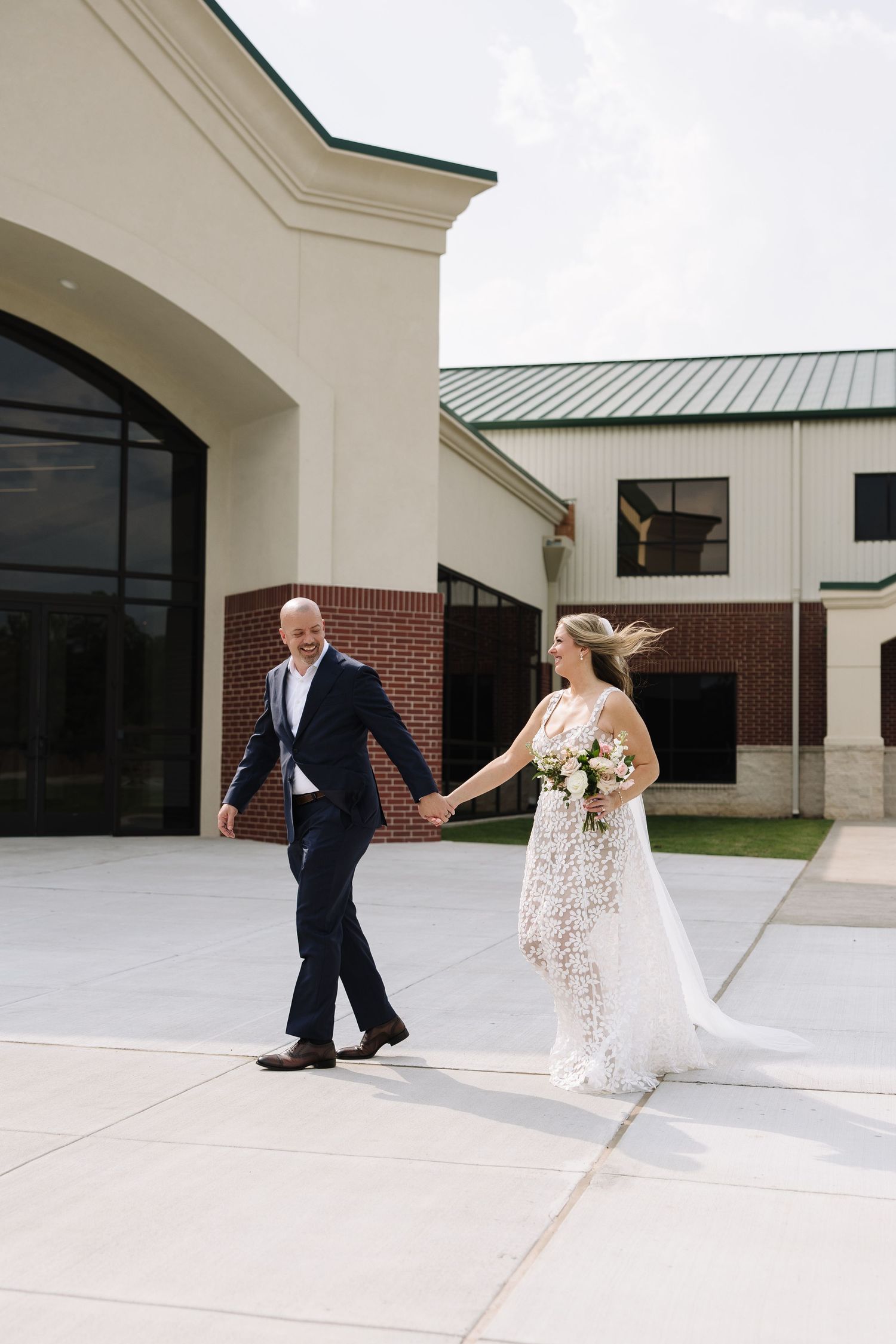 Bride and groom walk hand in hand outside a modern wedding venue, bride wearing lace gown holding bouquet.
