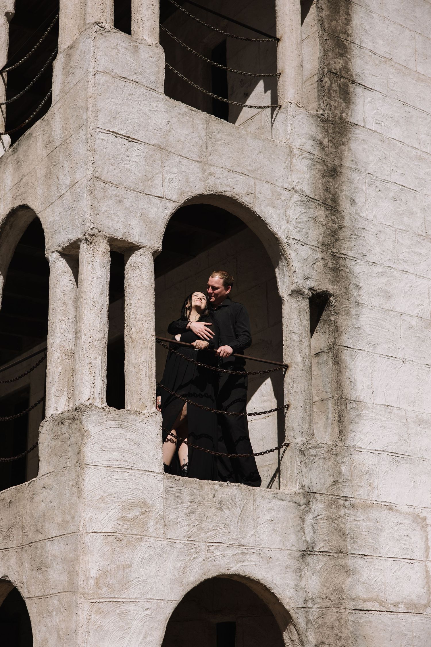 Couple in black embracing on a stone balcony of an ancient arched building with barbed wire railing.