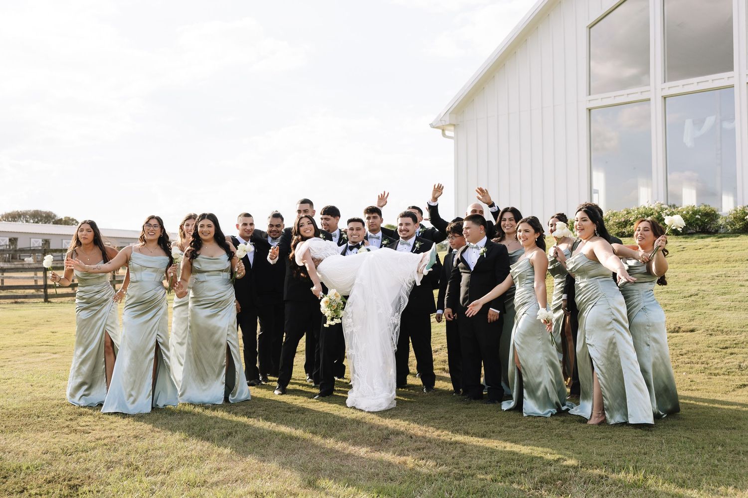 A wedding party poses together outside a white chapel on a sunny day.