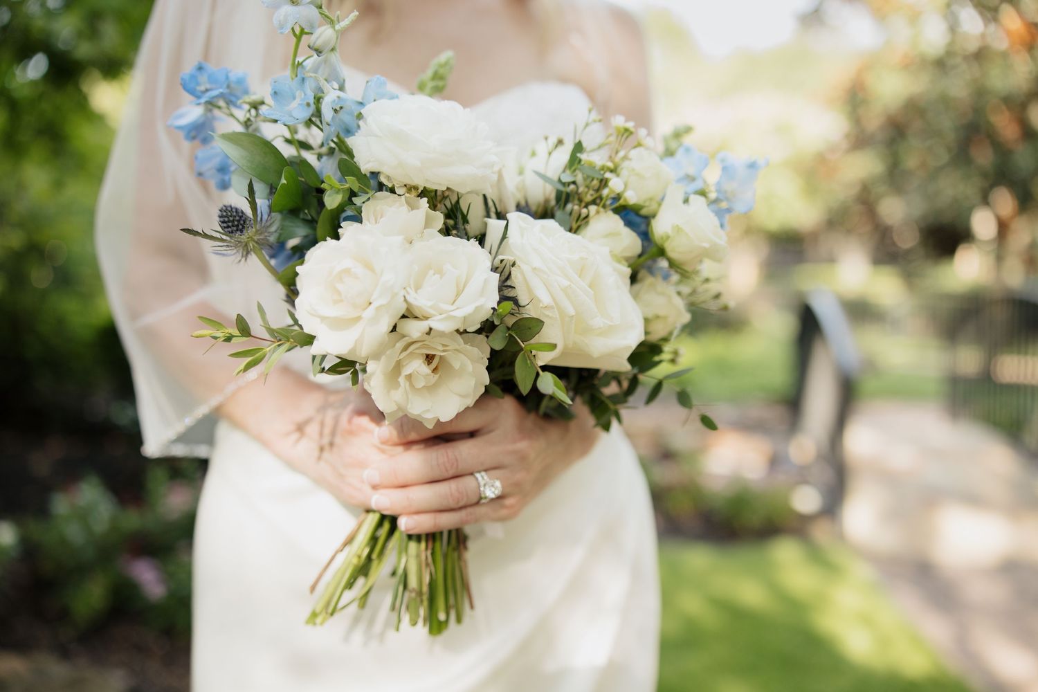 Delicate white and blue wedding bouquet with roses and greenery against a garden background.