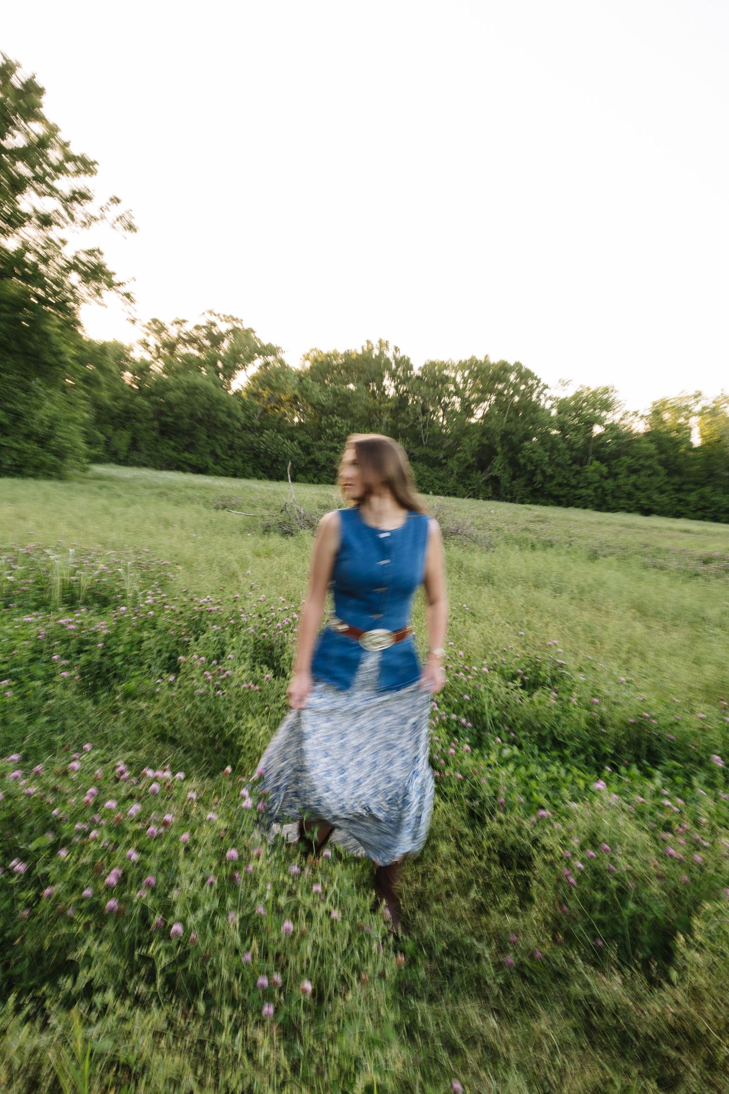 A woman in a blue top and floral skirt walks through a lush green meadow with wildflowers at sunset.