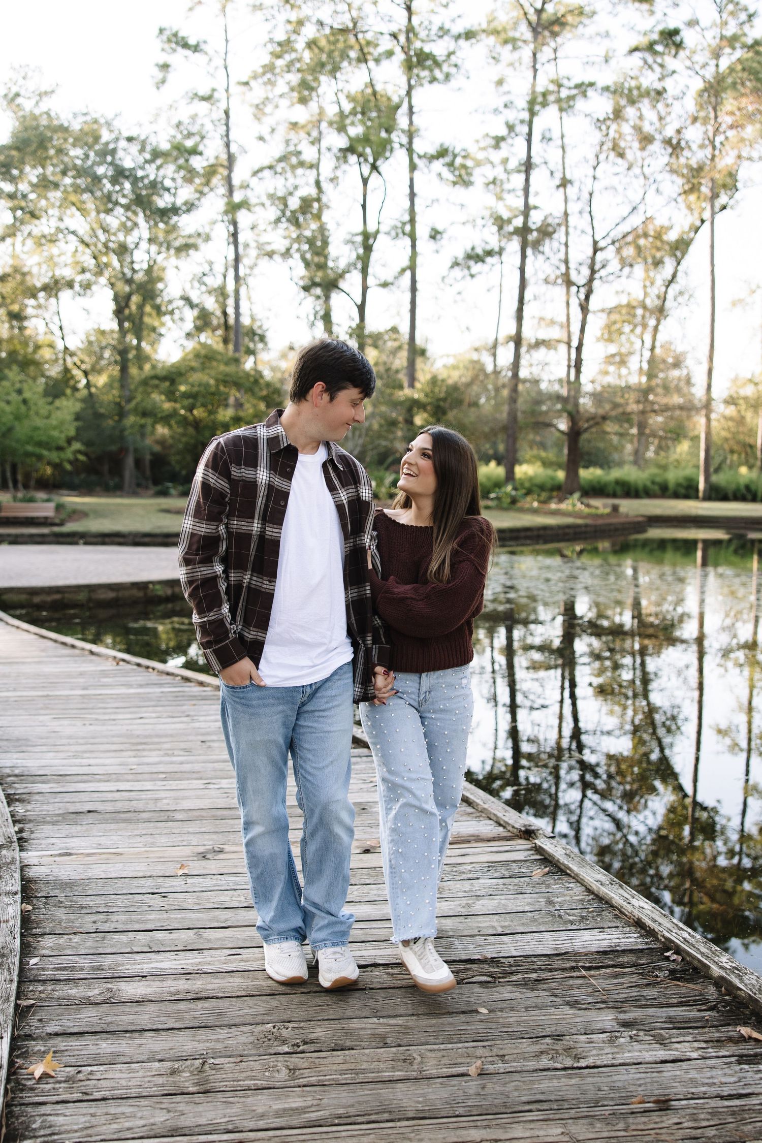 A couple walks together on a rustic wooden boardwalk over a tranquil pond surrounded by tall trees.