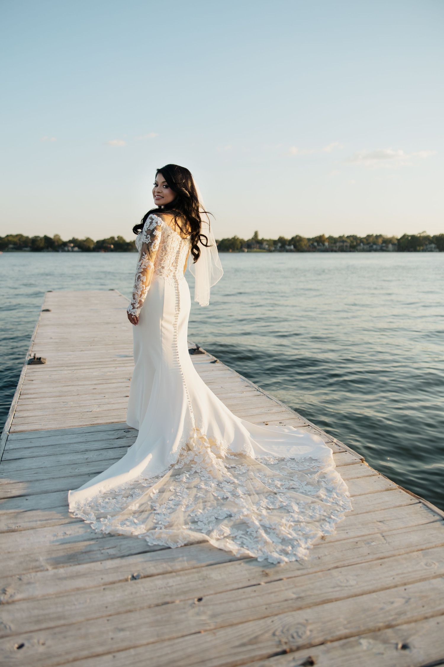 A bride in a flowing lace wedding dress poses on a wooden dock by a serene lake during sunset.