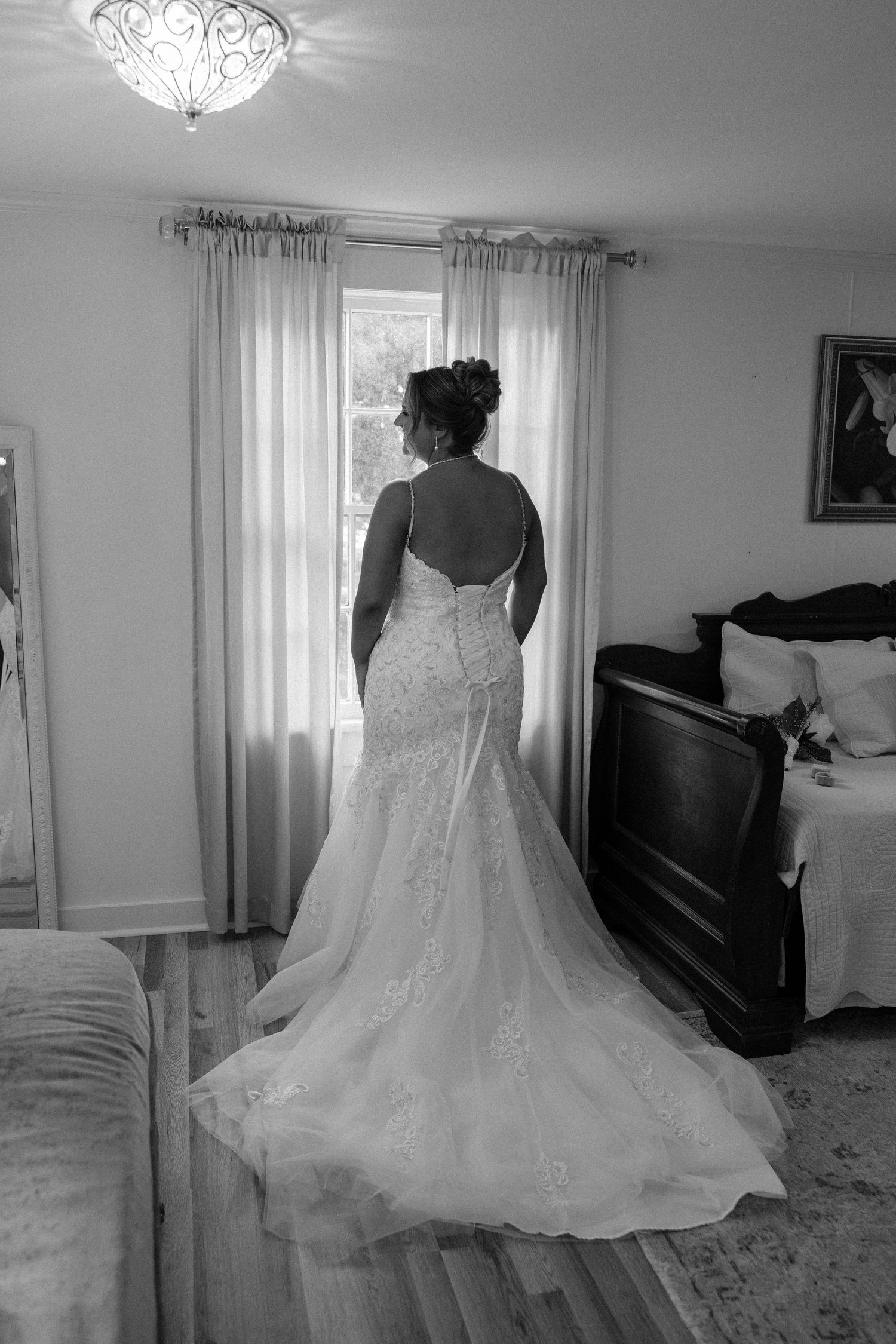 Black and white photo of a bride in a lace mermaid wedding gown with a train, standing before a sunlit window.