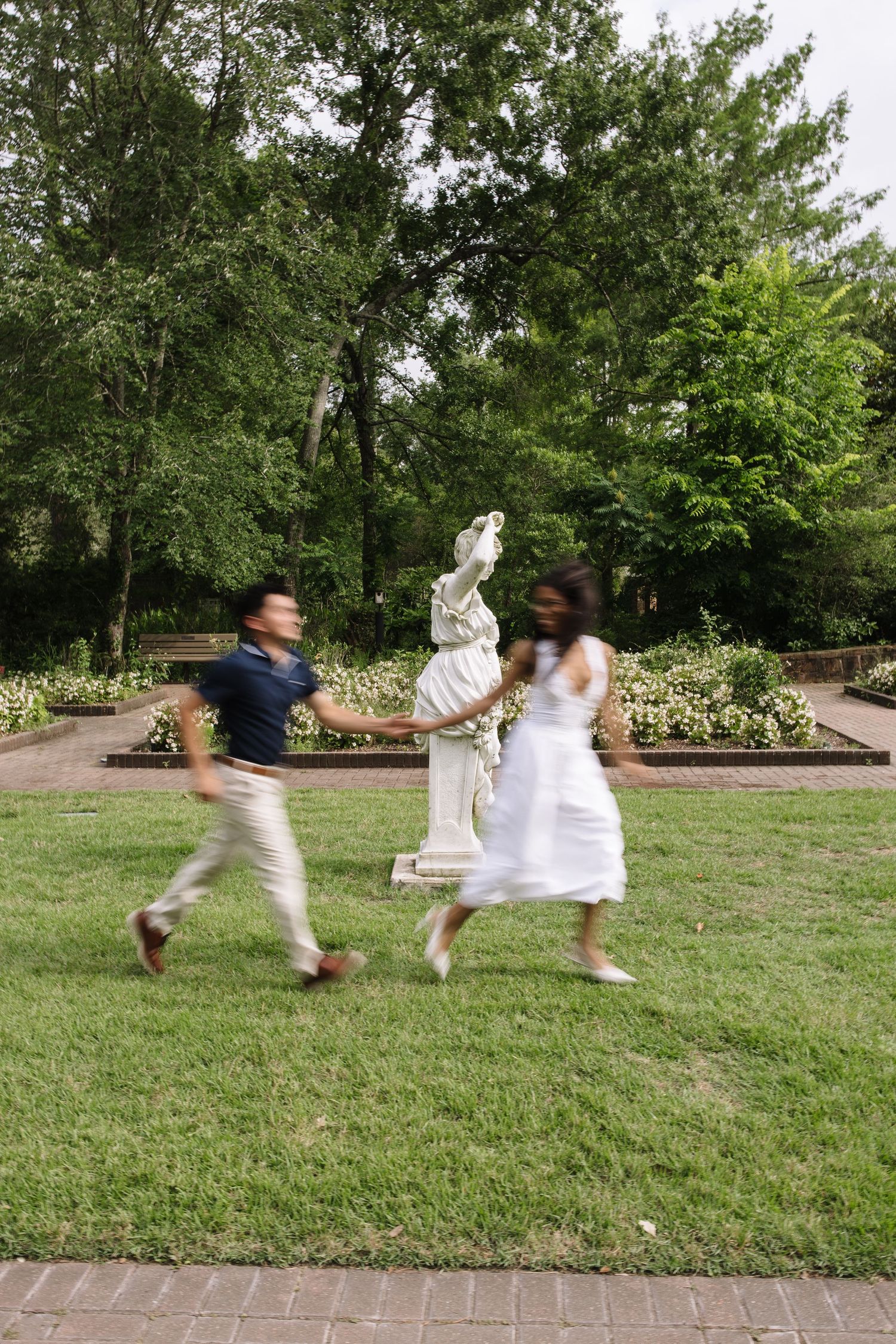Couple running hand-in-hand through a lush garden park with a white classical statue in the background.