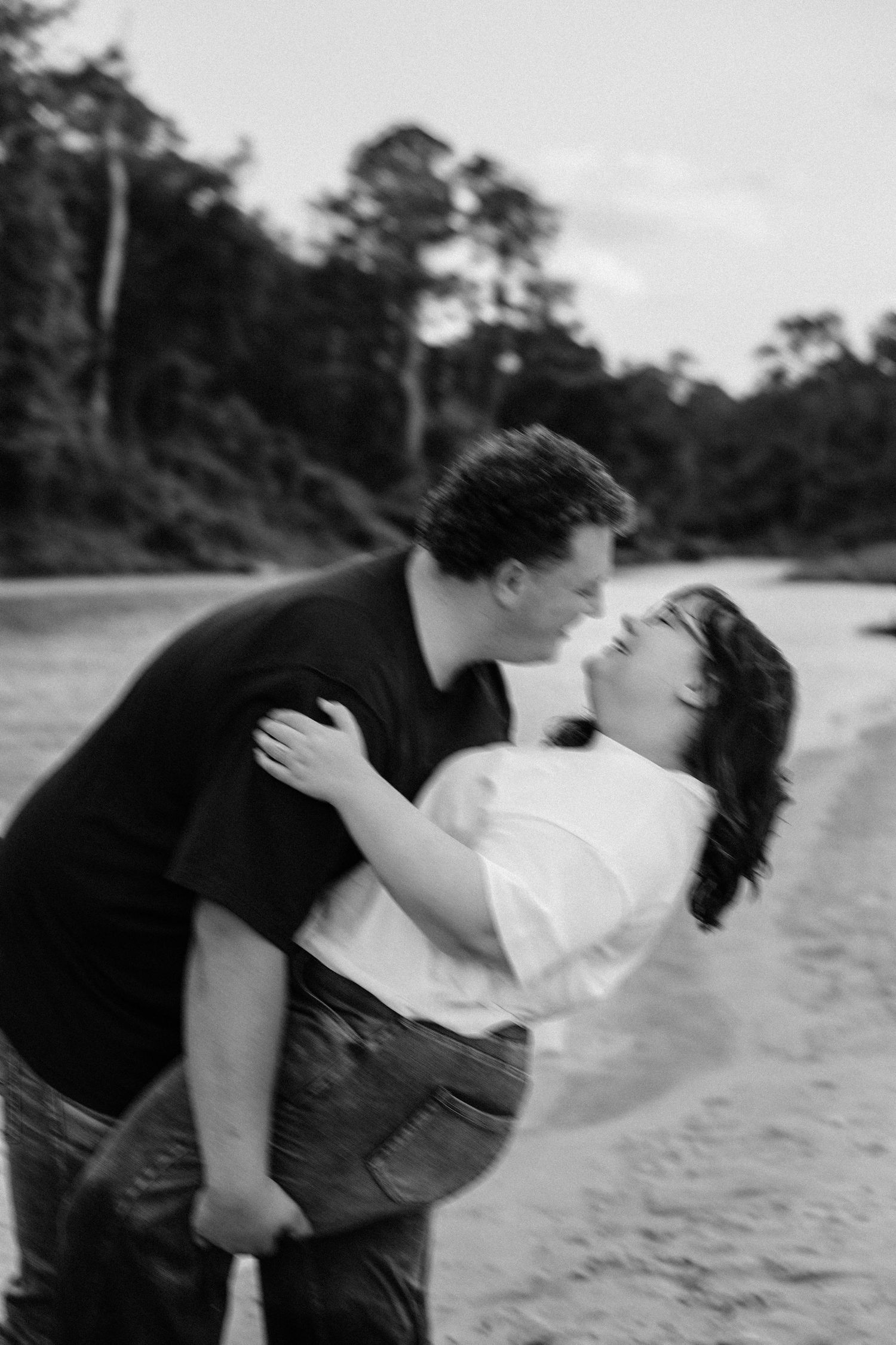Black and white photo of a couple sharing a romantic kiss on a beach surrounded by lush trees.