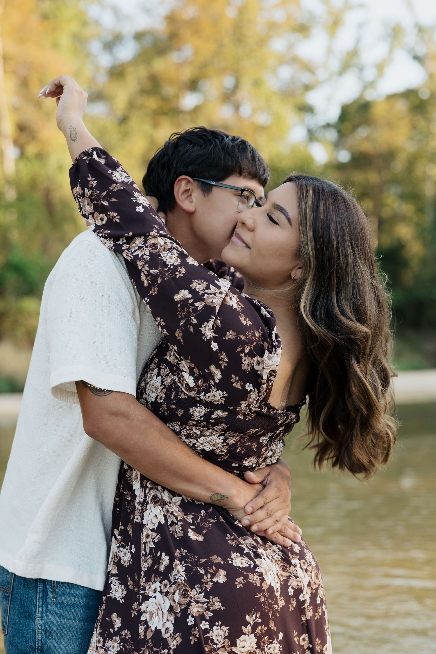 A romantic couple embraces by a lake, with the person in a brown floral dress and the other in a white shirt and jeans.