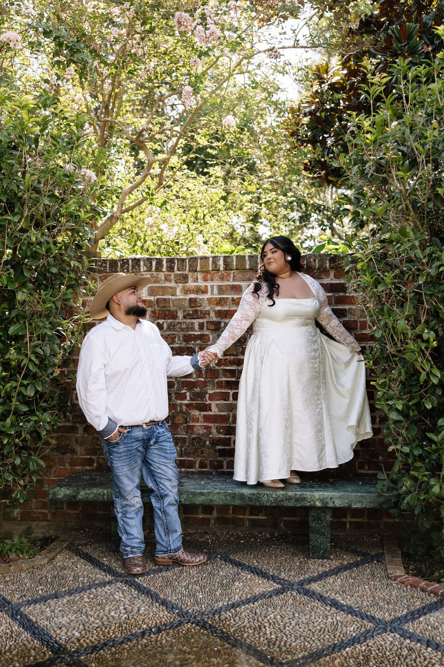 Bride in white lace gown stands on bench holding hands with groom in cowboy hat and jeans in a garden.