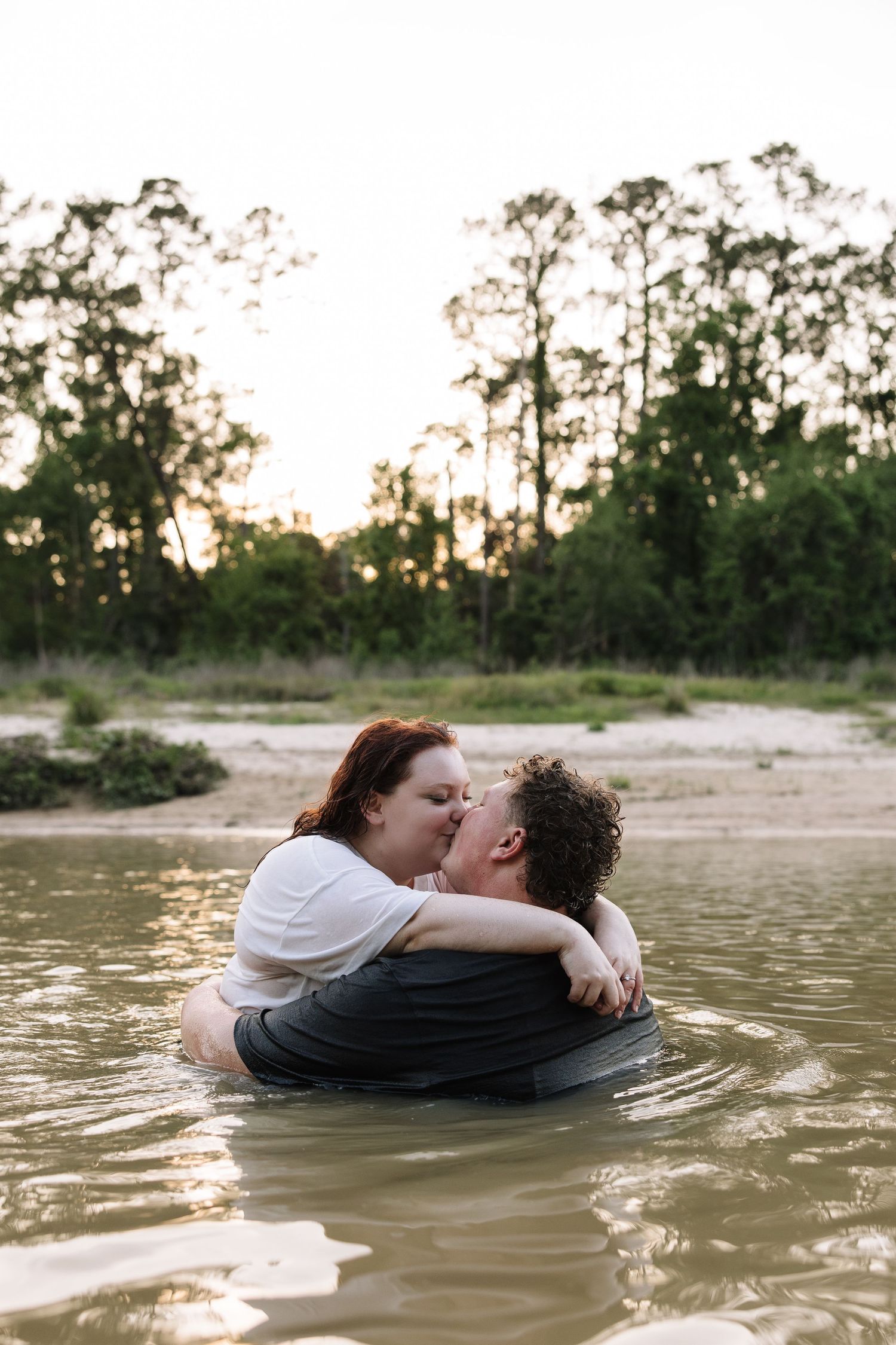 Couple sharing a romantic kiss while standing in a shallow river surrounded by trees at golden hour.