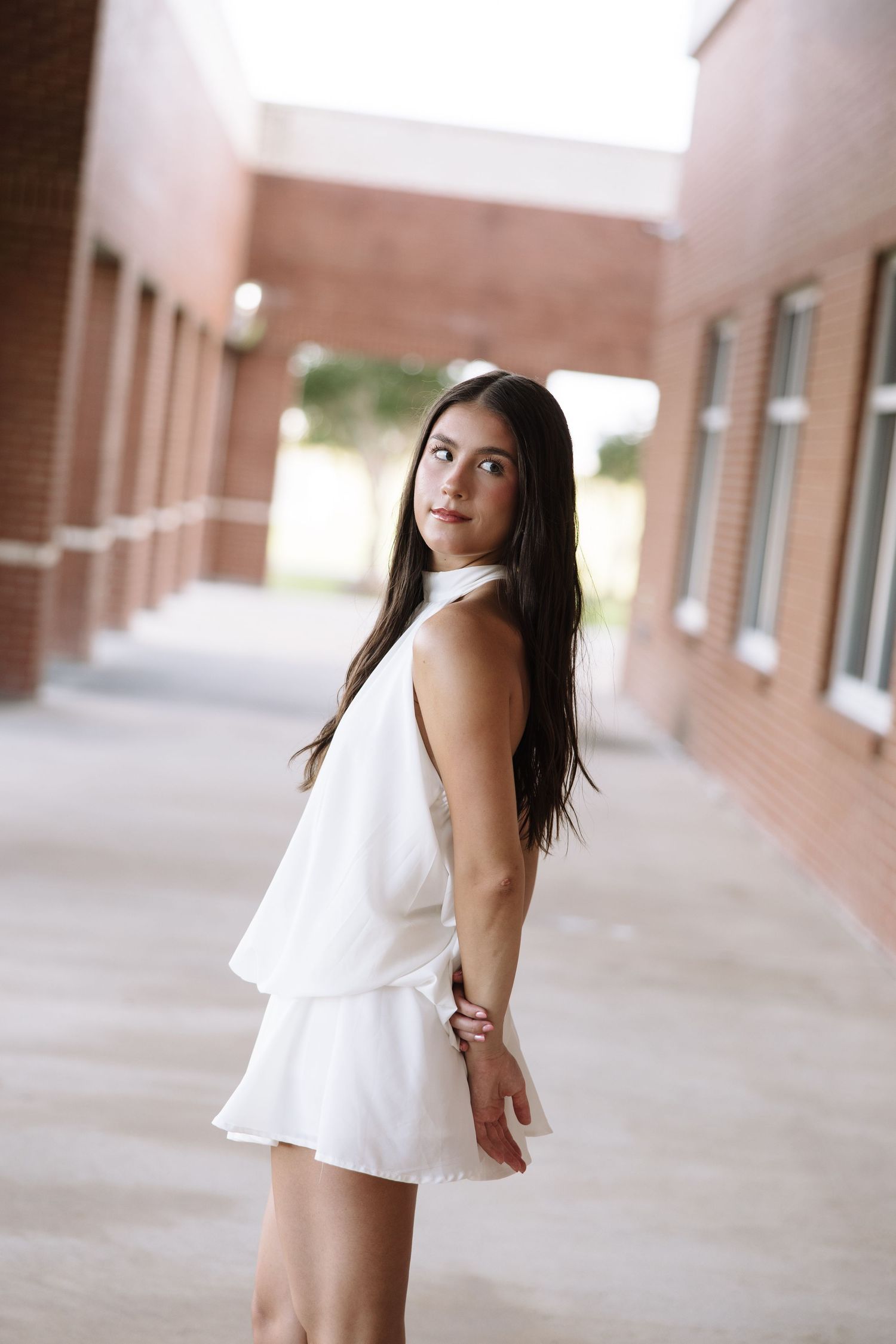 Young woman in white dress posing outdoors near brick building, long dark hair, looking over shoulder.