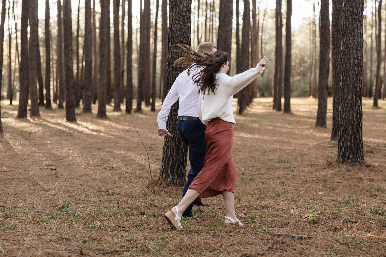 Two people dancing together in a pine forest with long shadows cast by tall trees in warm sunlight.