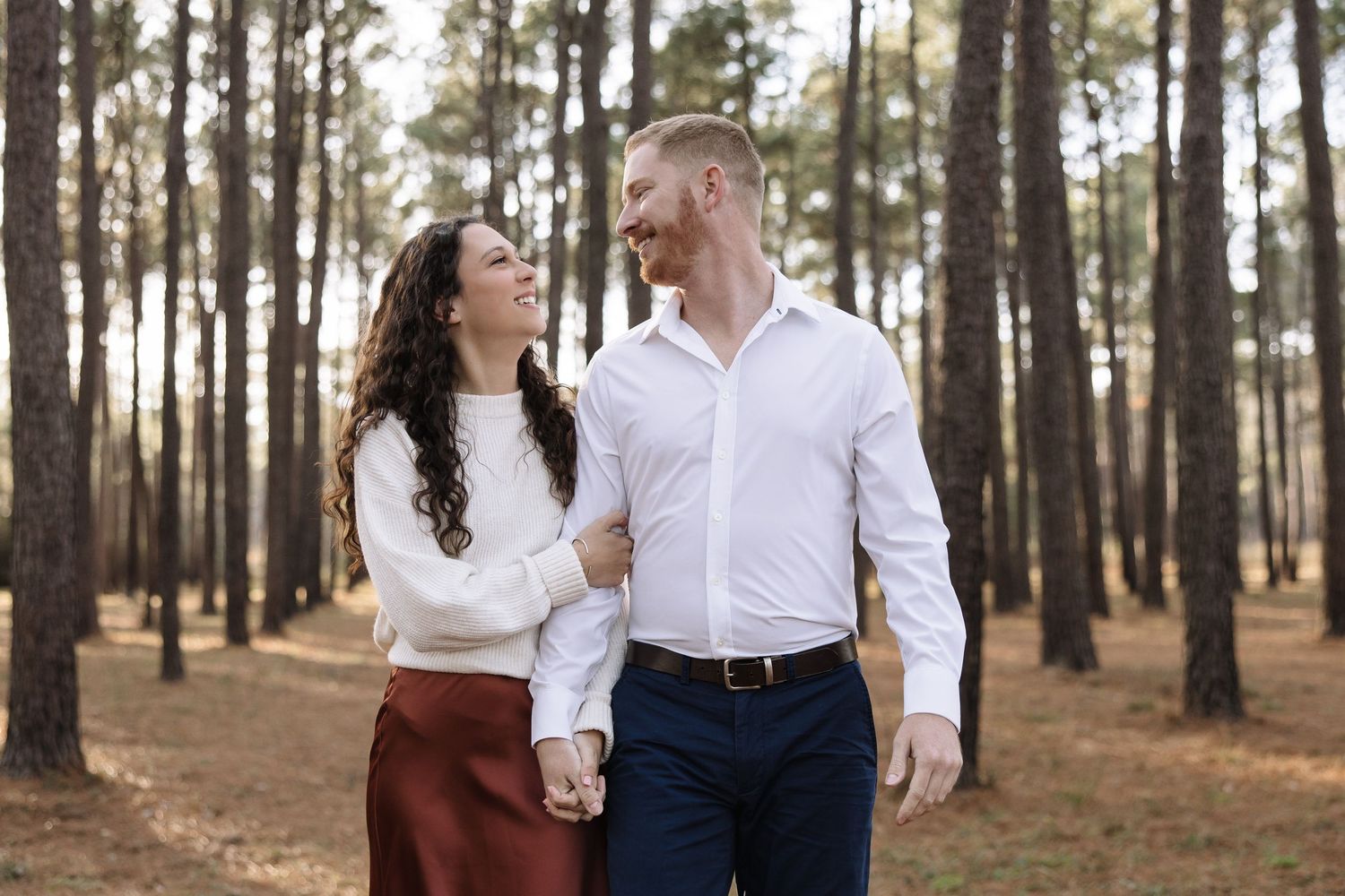 A couple in white tops and dark bottoms laughs together while walking through a serene pine forest.