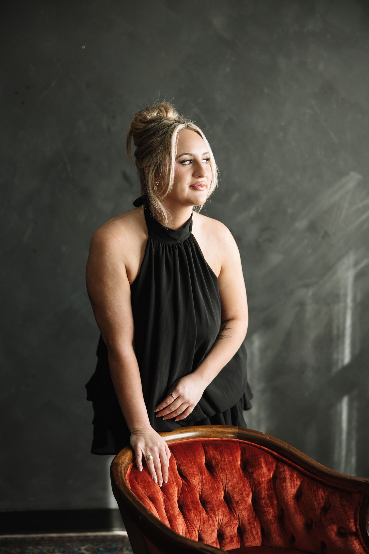 A moody portrait session against a dark background featuring a black sleeveless top and red velvet chaise lounge.