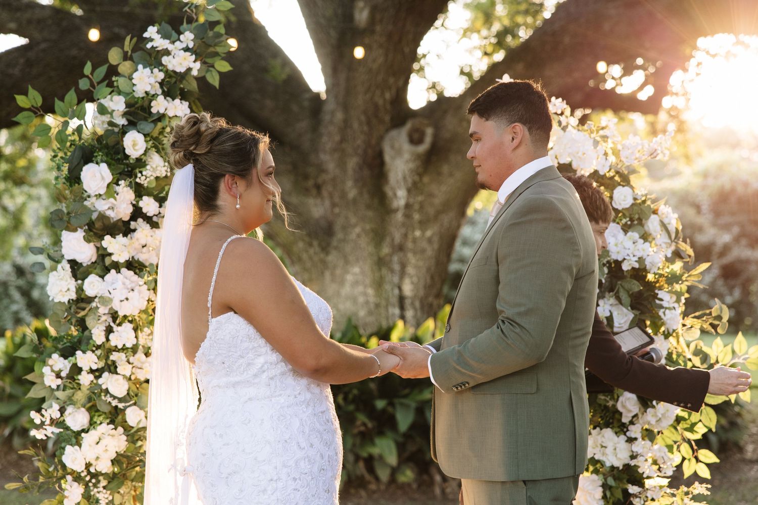 Bride and groom holding hands at floral arch ceremony outdoors under a tree with white roses.