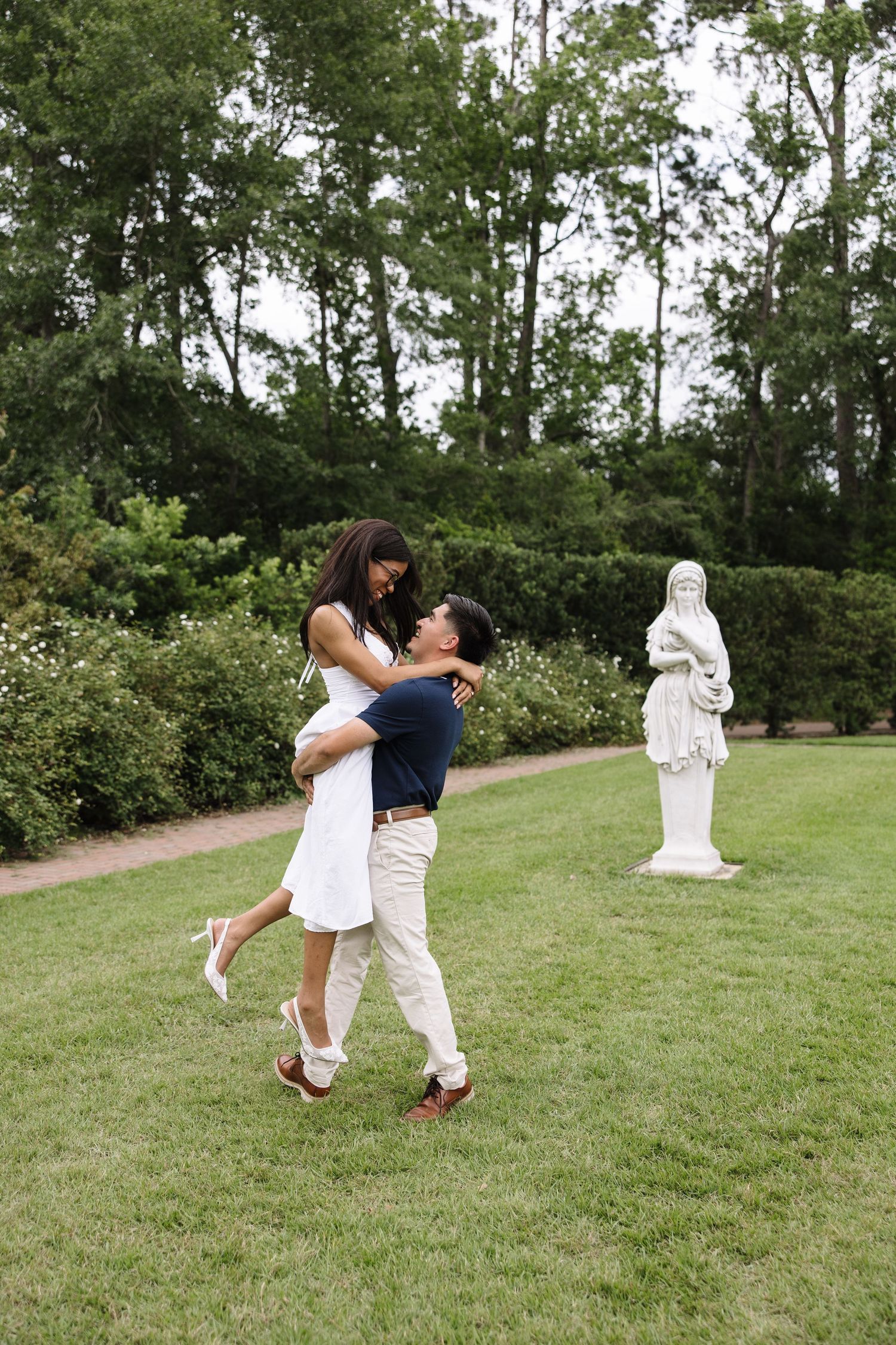Couple embracing in a lush garden, man lifting woman in white dress near a classical statue.