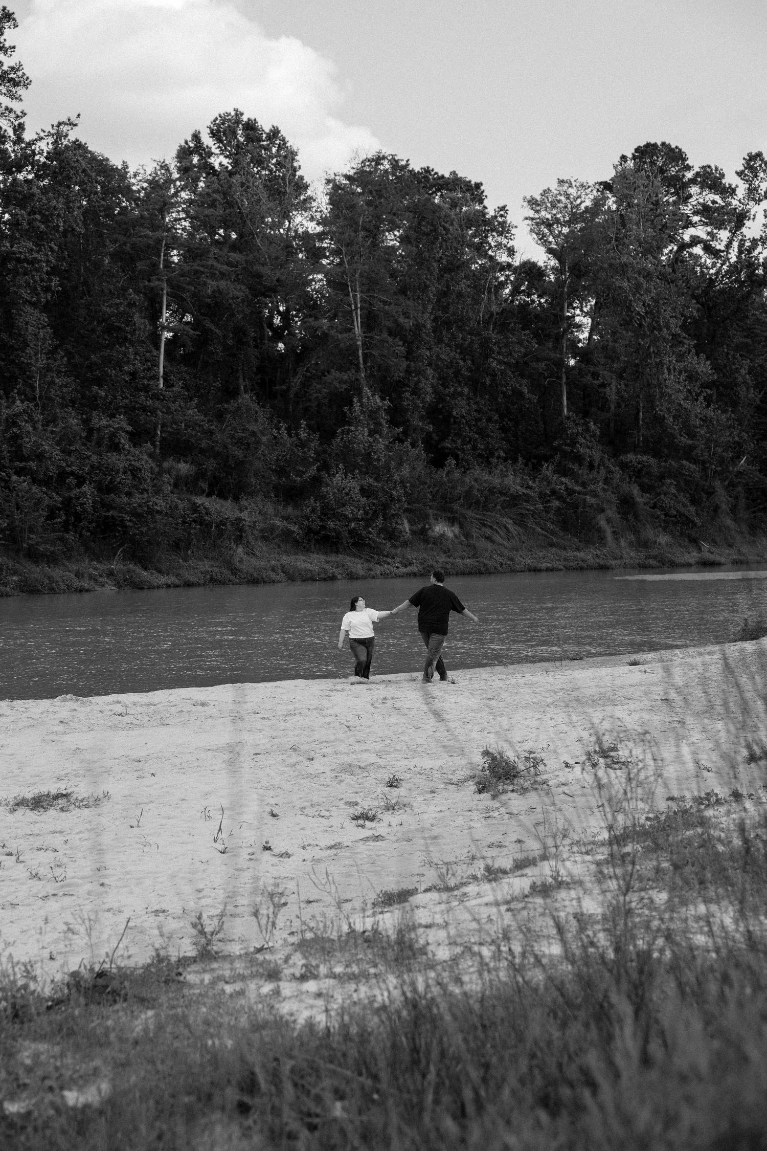 Black and white photo of a couple holding hands walking along a sandy riverbank surrounded by lush trees.