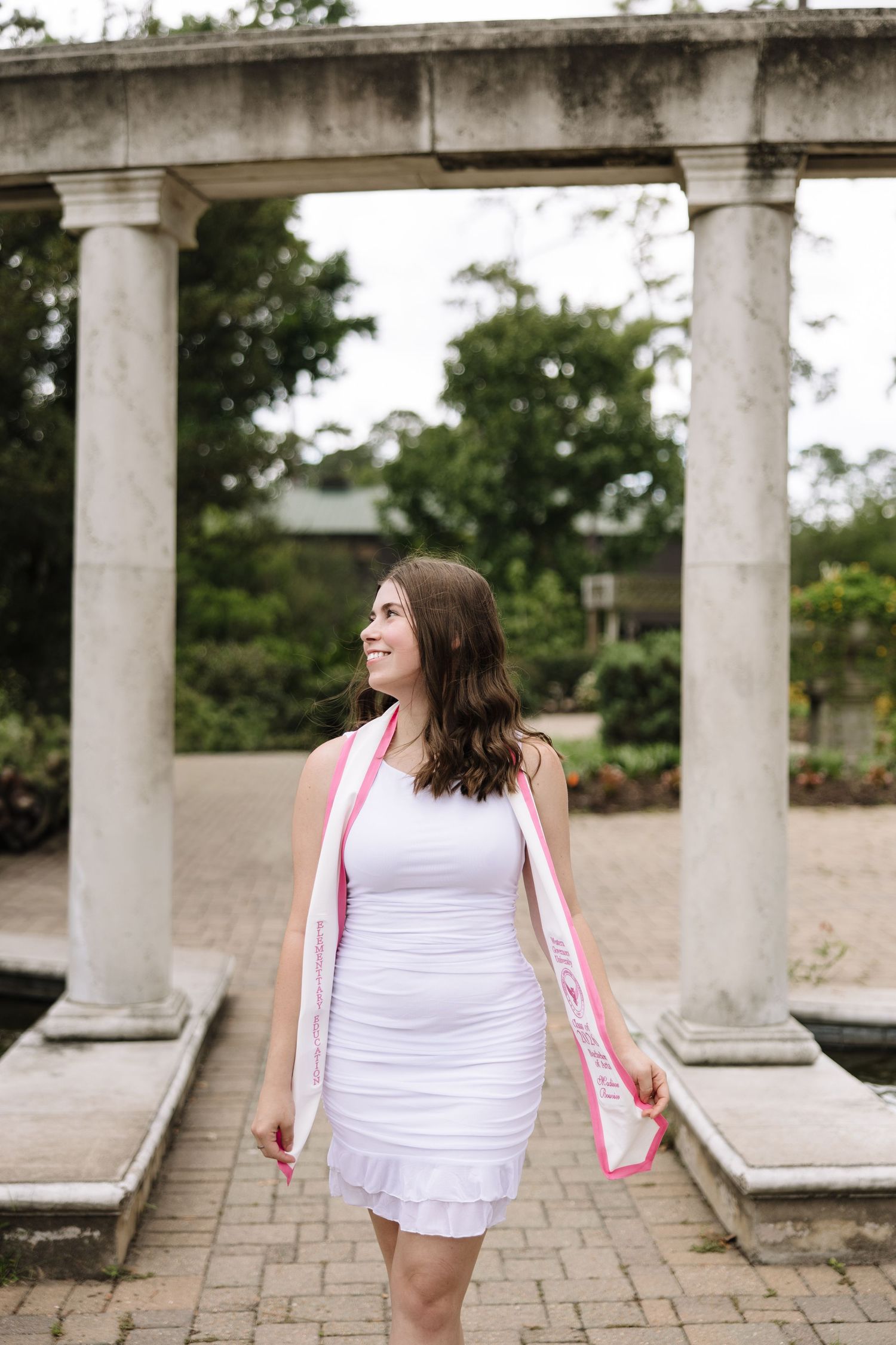 Young woman in white dress with pink sash smiles among stone columns in a garden setting.