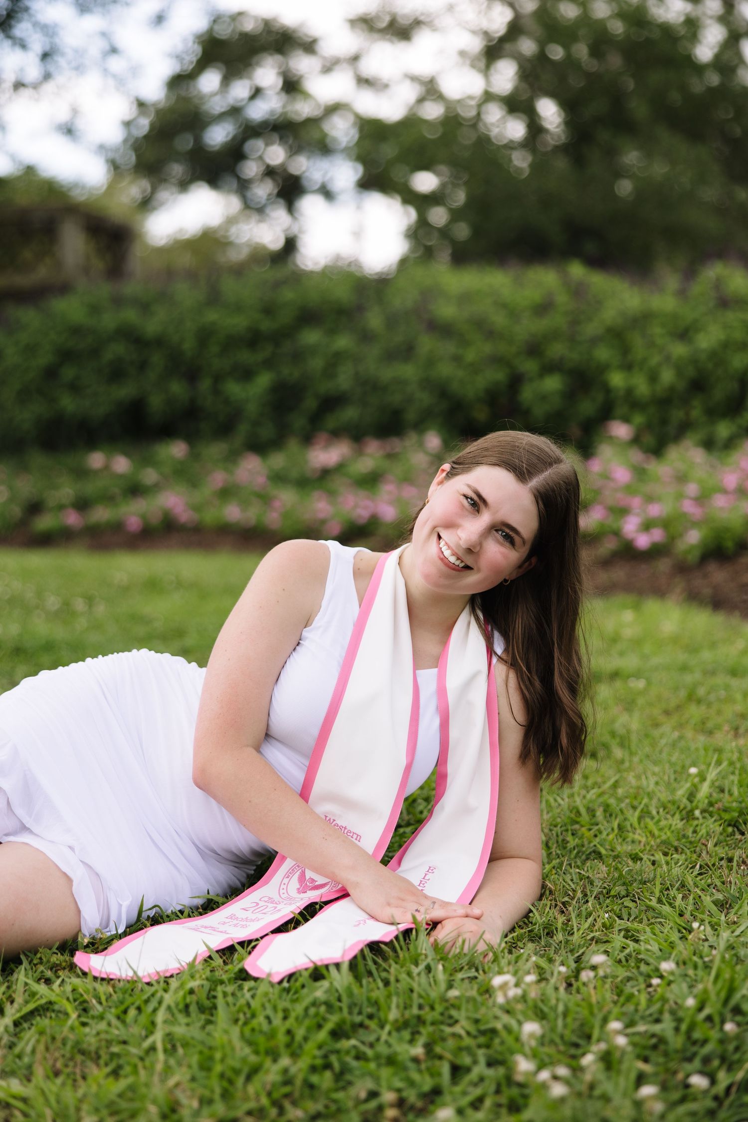 Young woman in white dress and pink scarf smiling while relaxing on green grass in a blooming garden.
