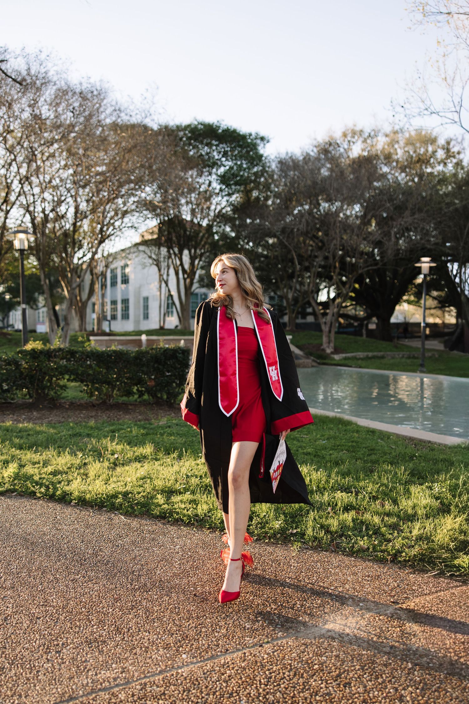 Young woman in black graduation gown and red dress walks confidently on campus path near a pond.