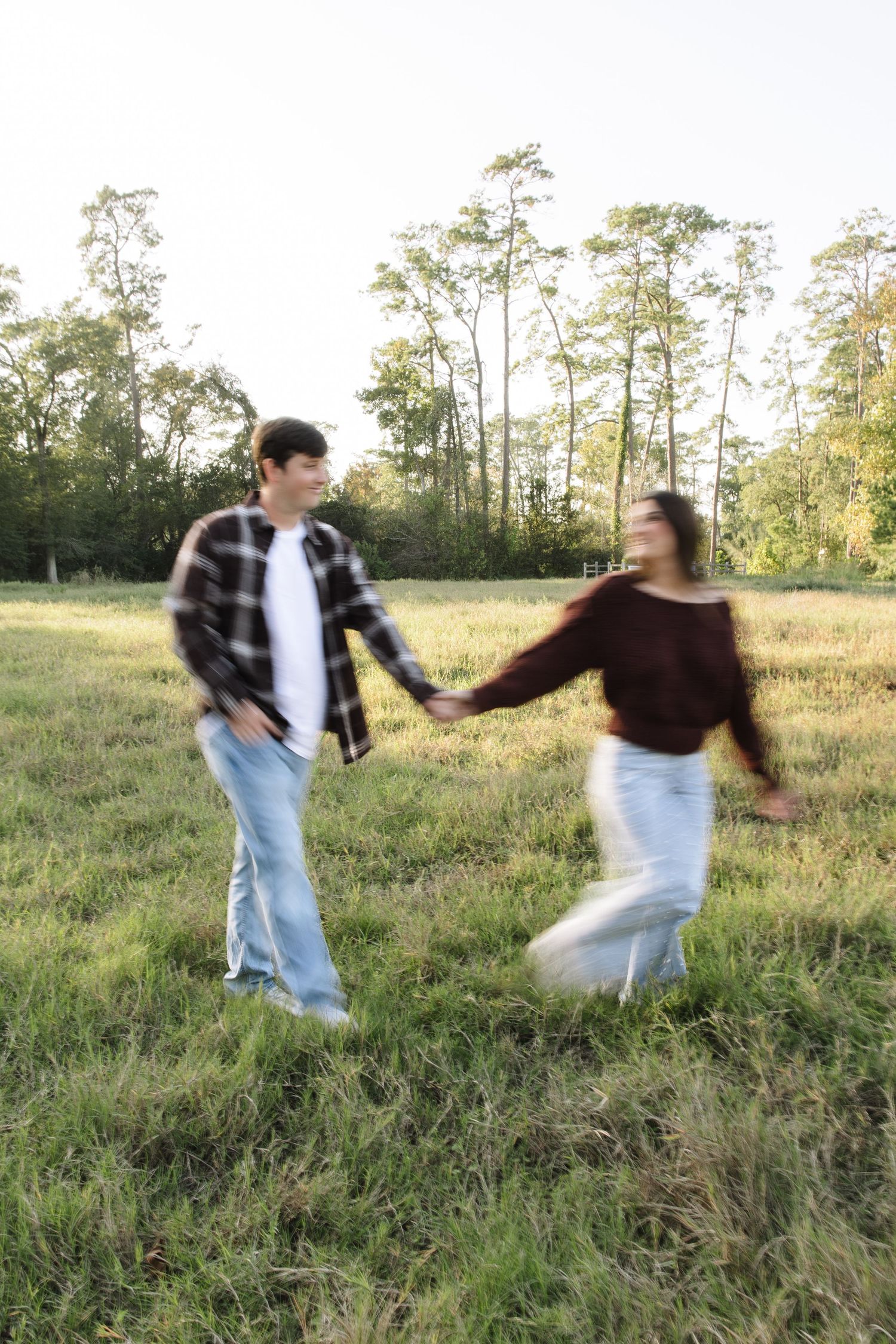 A couple spins playfully in a grassy field at sunset while wearing casual denim and dark clothing.