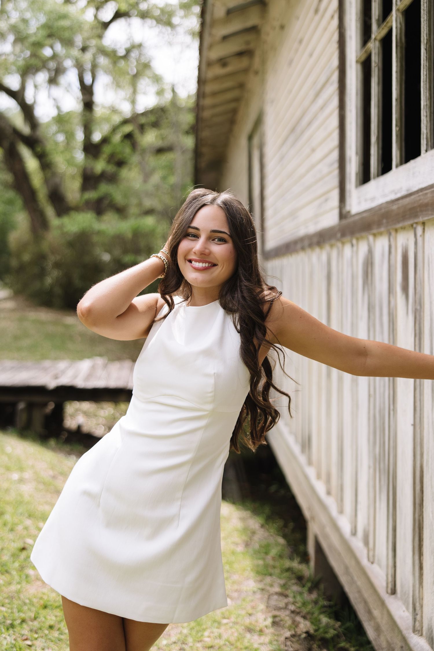 Young woman in white mini dress smiling outdoors beside rustic building with lush green trees in background.
