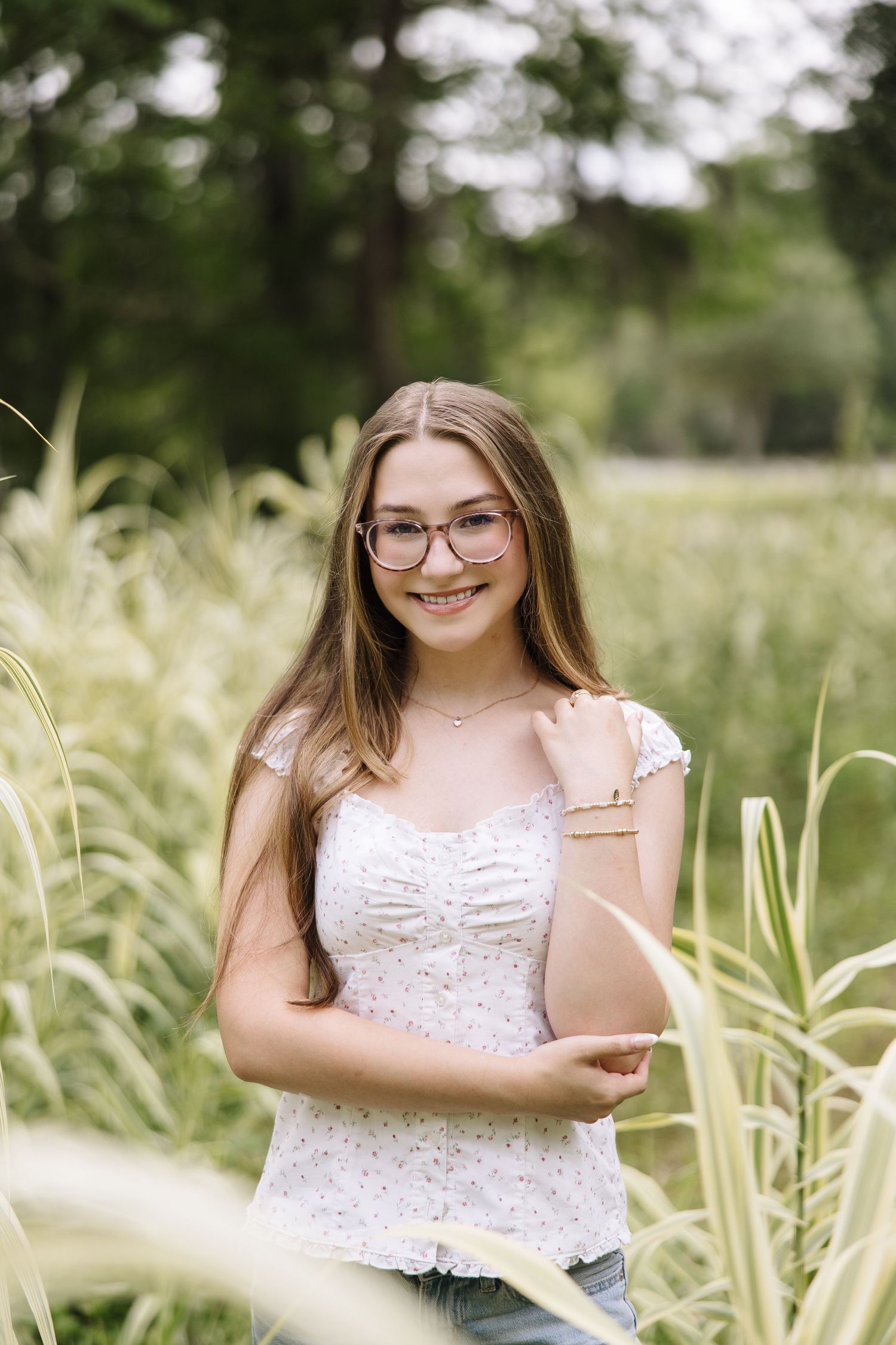 Young woman with glasses and long hair smiles in a white floral top, standing among tall green grasses outdoors.