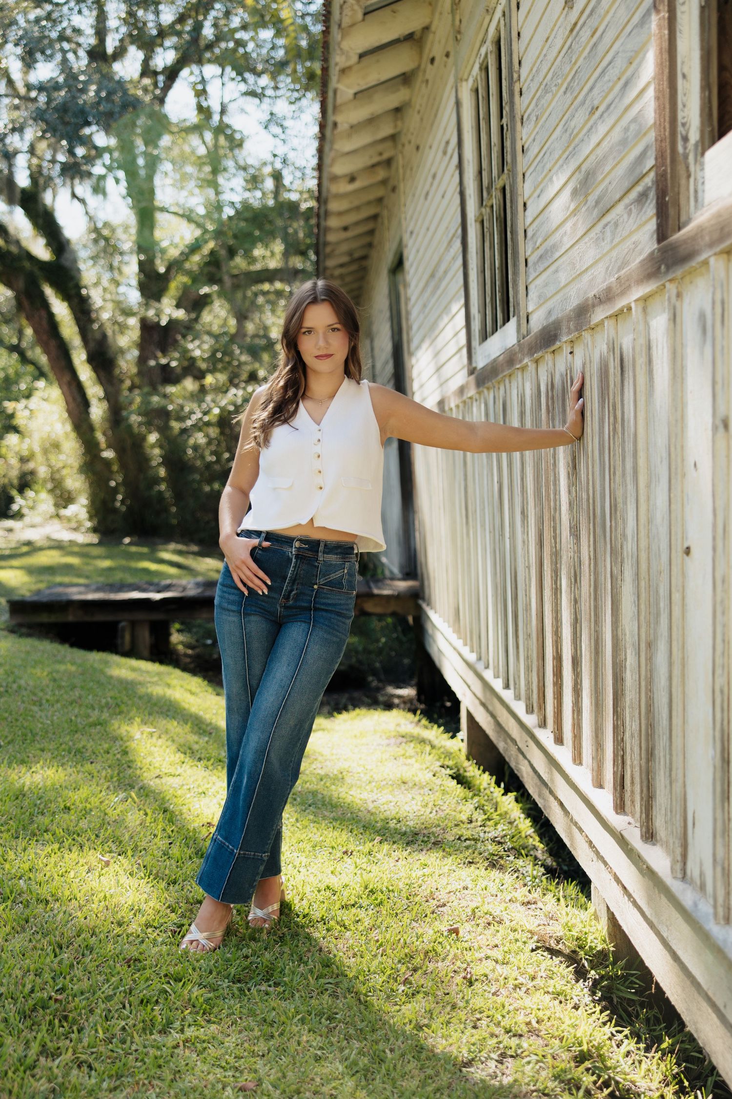 Woman in white blouse and jeans poses against rustic wooden wall in outdoor sunlit setting.