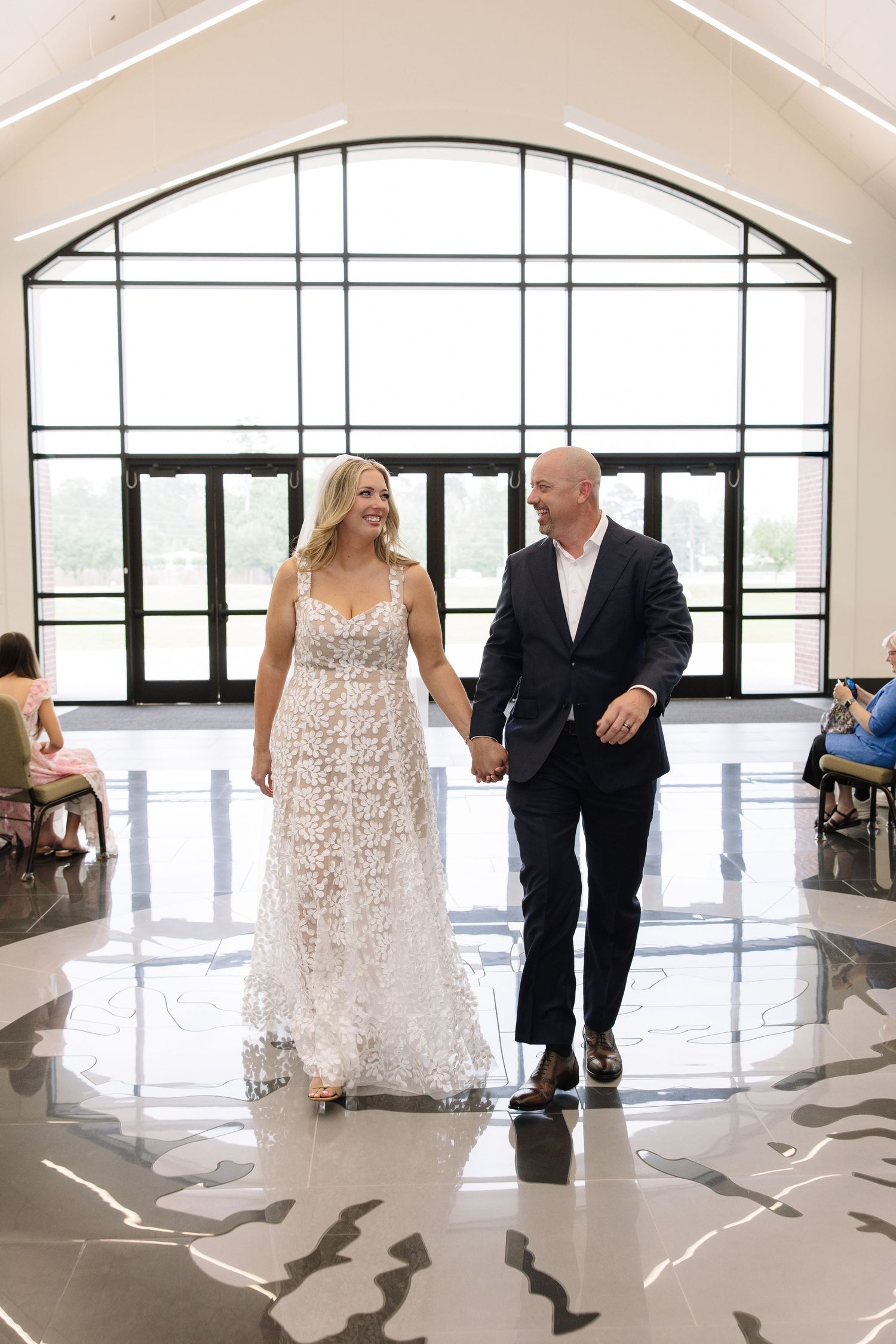 Bride in lace gown and groom in navy suit walk hand-in-hand through elegant venue with arched windows.