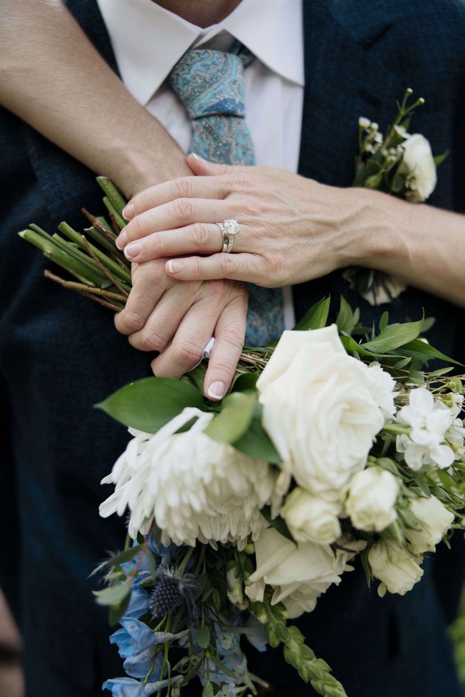 Close-up of hands with wedding ring and white rose bouquet with blue accents.