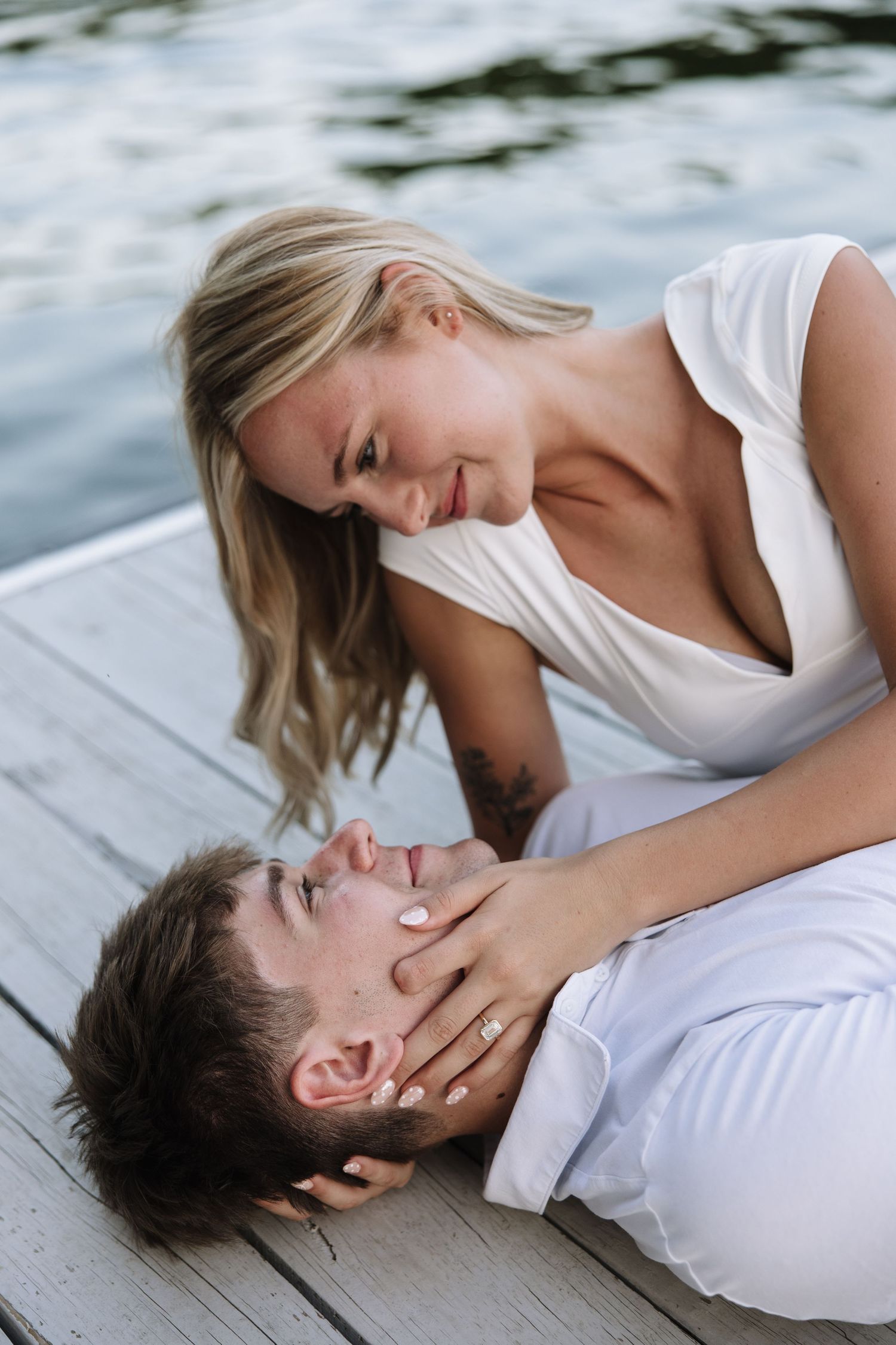 Couple in white sharing a romantic moment on a wooden dock by the water, woman smiling down at man lying in her lap.