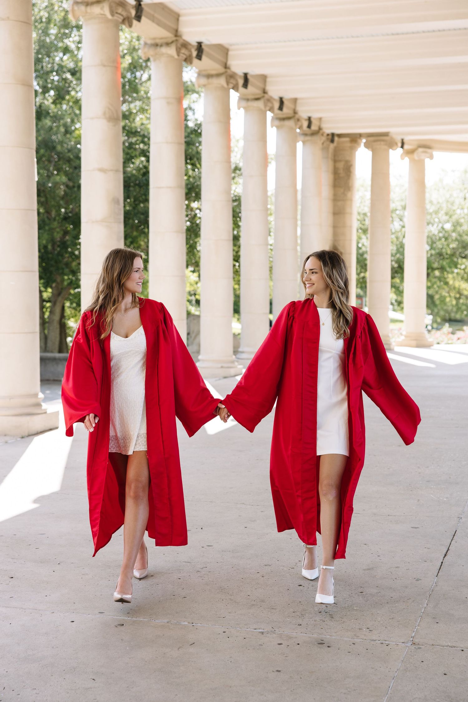 Two women in red graduation gowns and white dresses hold hands walking through a columned corridor.