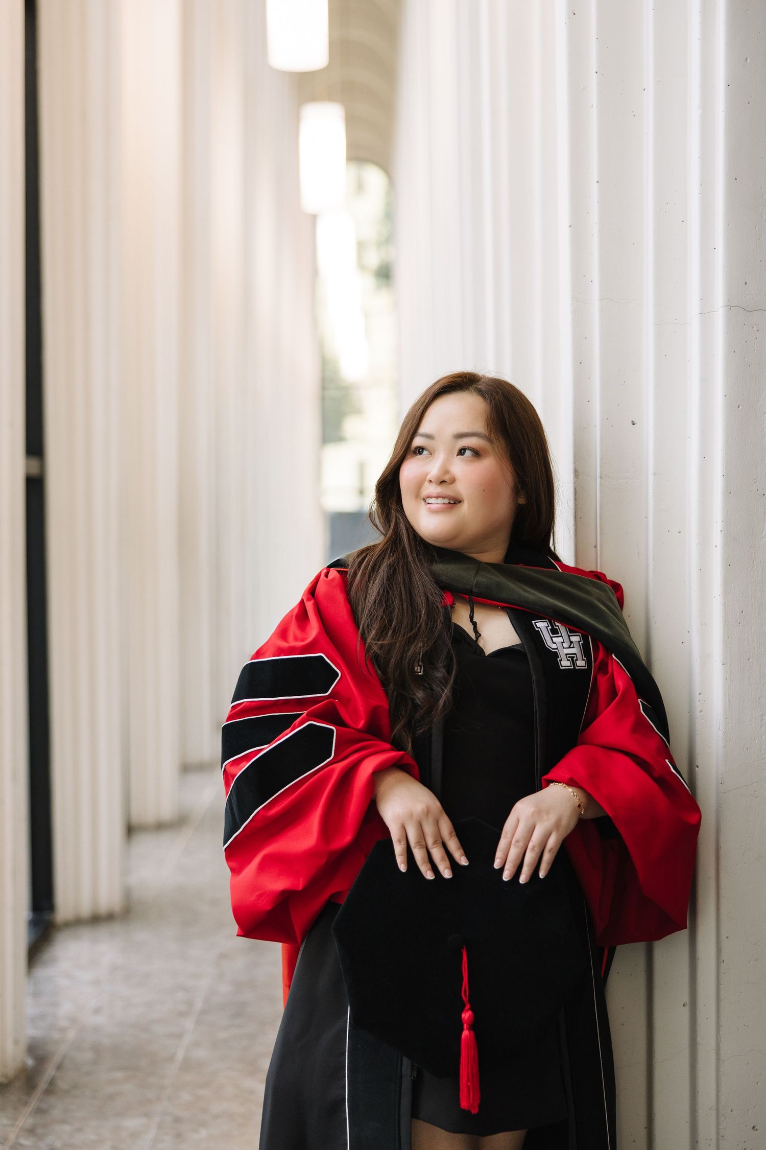 A smiling woman in red and black doctoral graduation regalia leans against a white column outdoors.