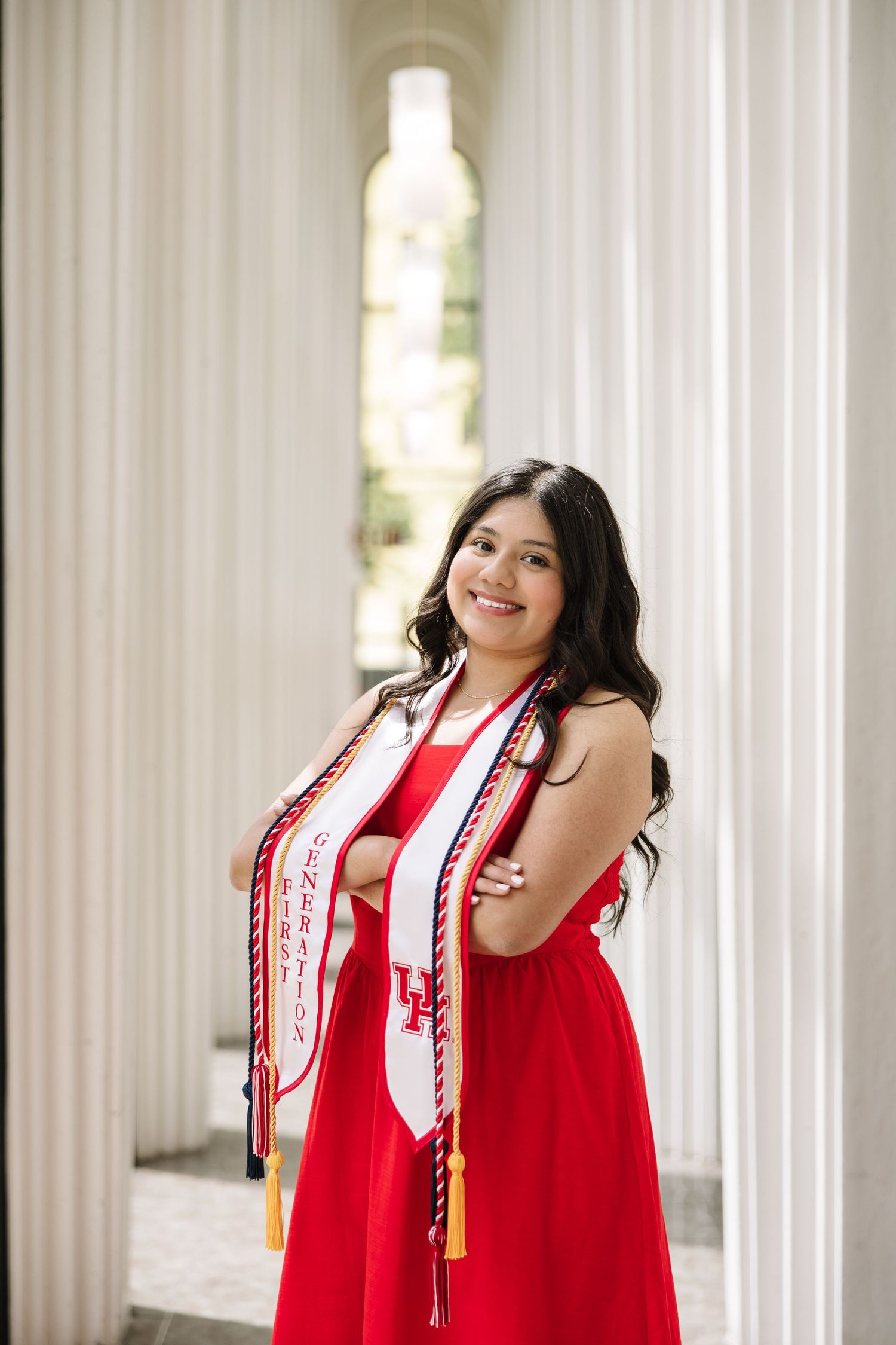 Smiling graduate in red dress with University of Houston stoles and honor cords poses between white columns.