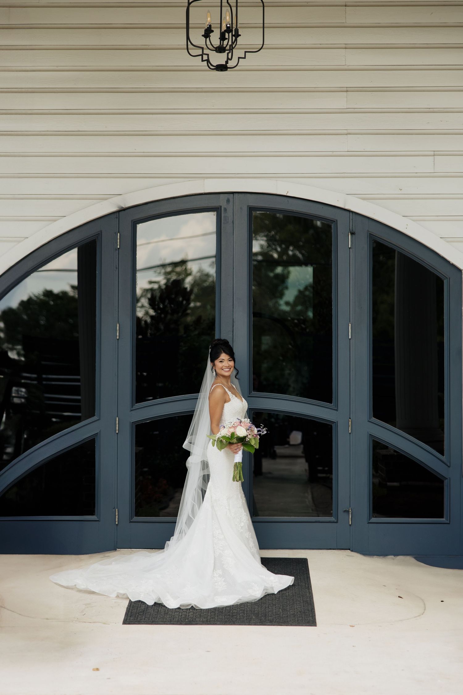 A bride in a white wedding dress poses elegantly against dark arched windows on a porch.