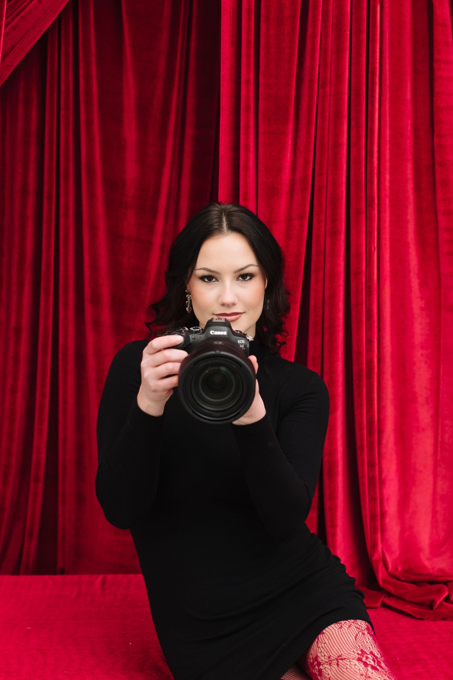 A photographer in black clothing poses with a camera against a vibrant red curtain backdrop.