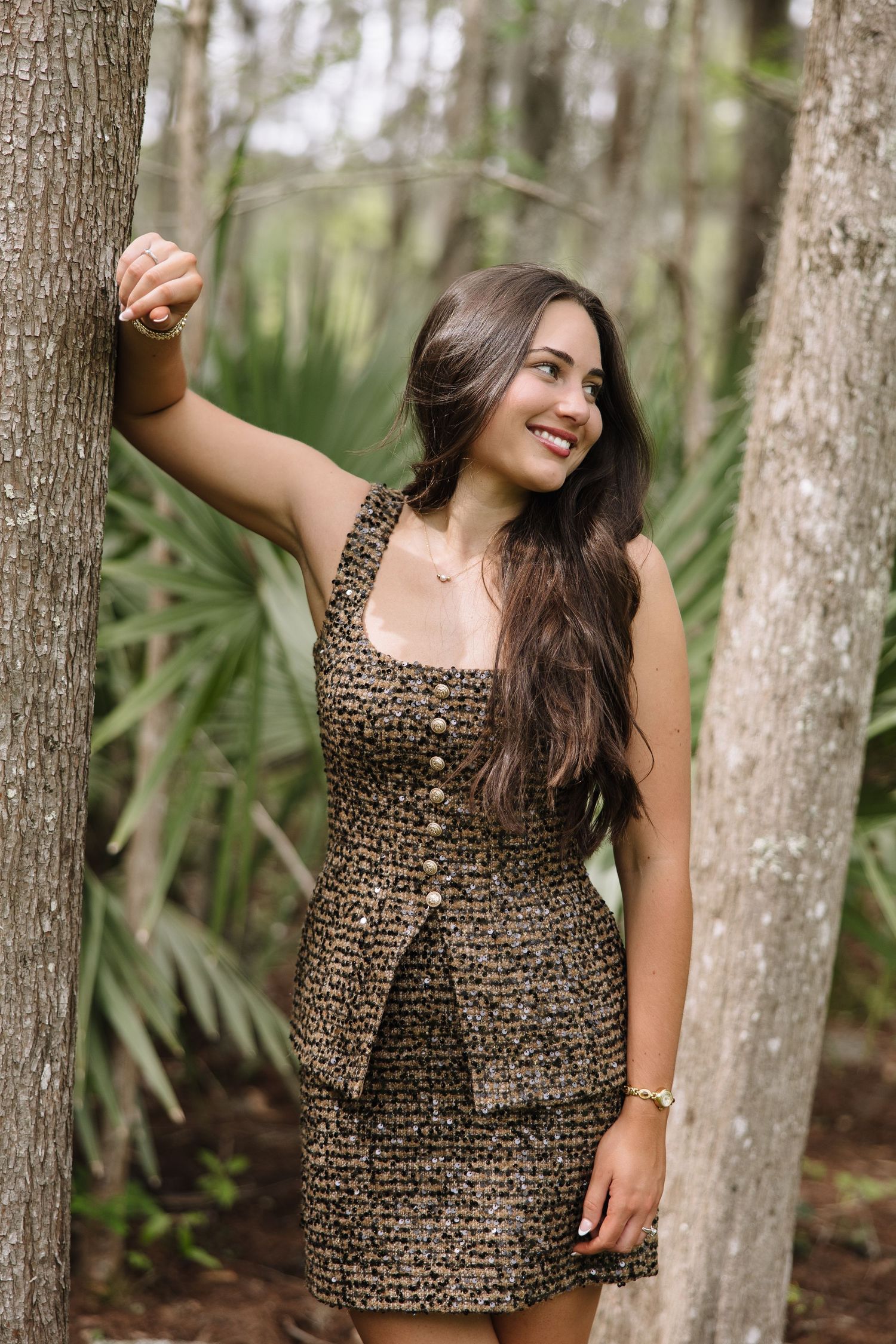 Smiling brunette woman in leopard print dress posing between trees in a lush green forest setting.