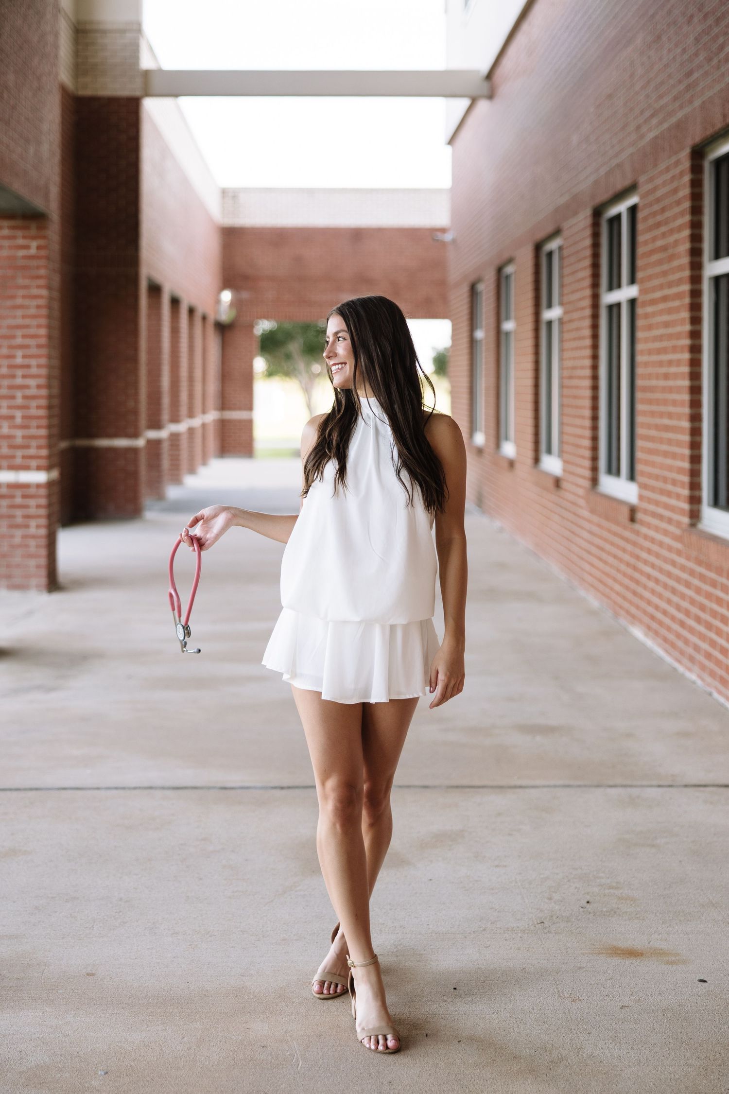 Young woman in white halter dress and sandals smiling while walking in a brick building corridor.