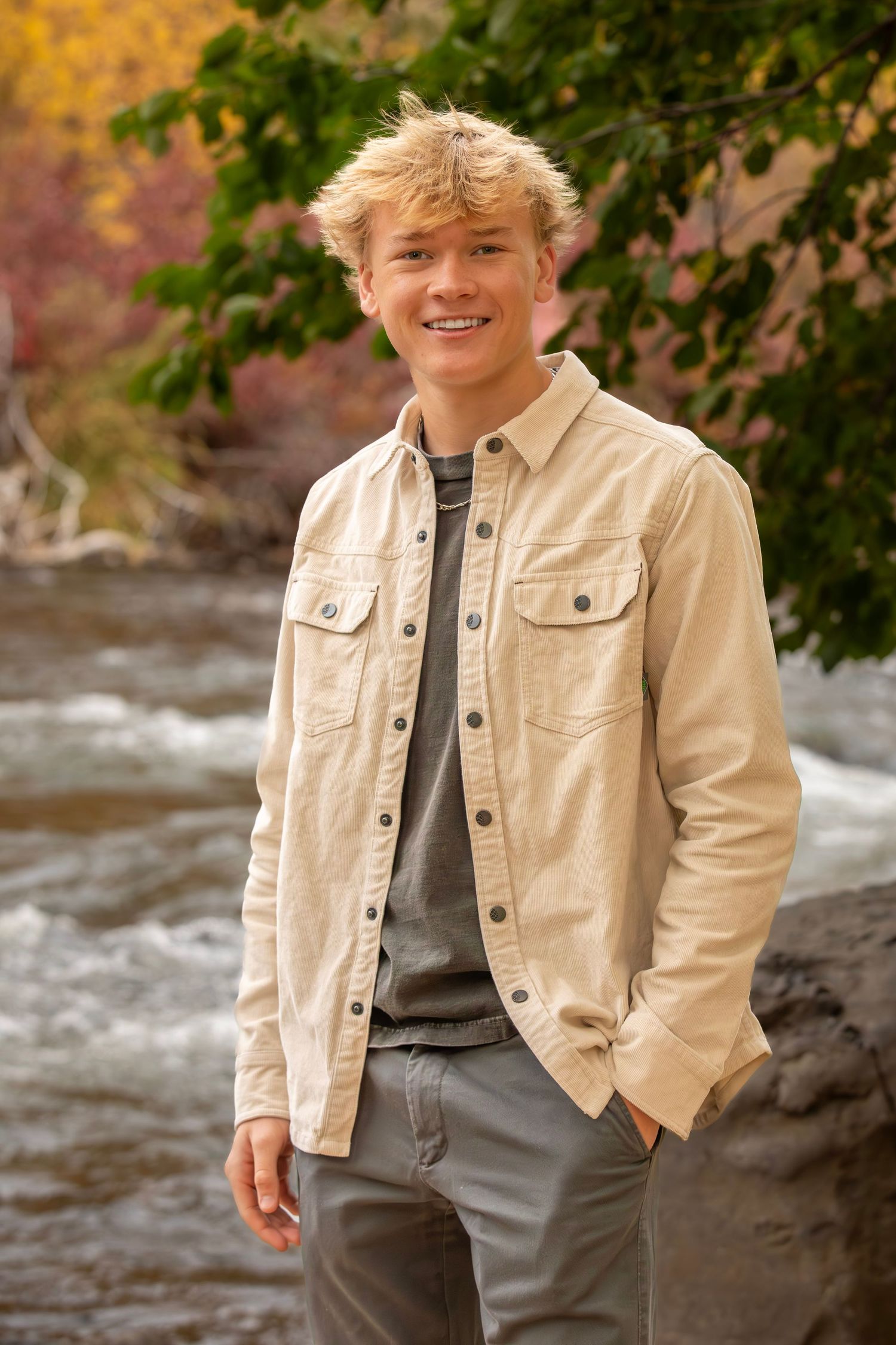 A person in a beige jacket and gray t-shirt stands smiling by a rushing river with autumn foliage in the background.