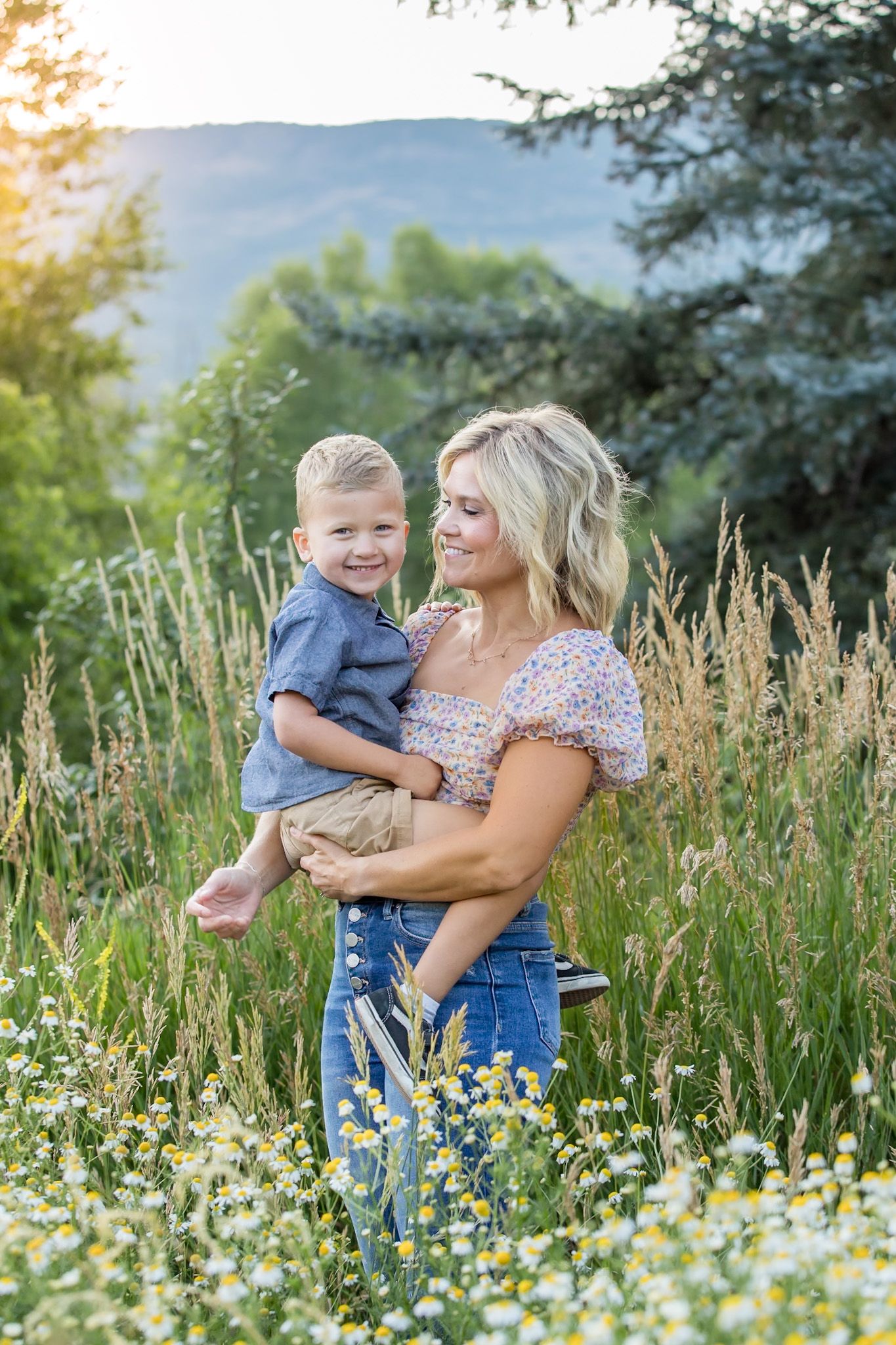 Woman in floral top holding young child while standing in sunny wildflower meadow with mountains in background.