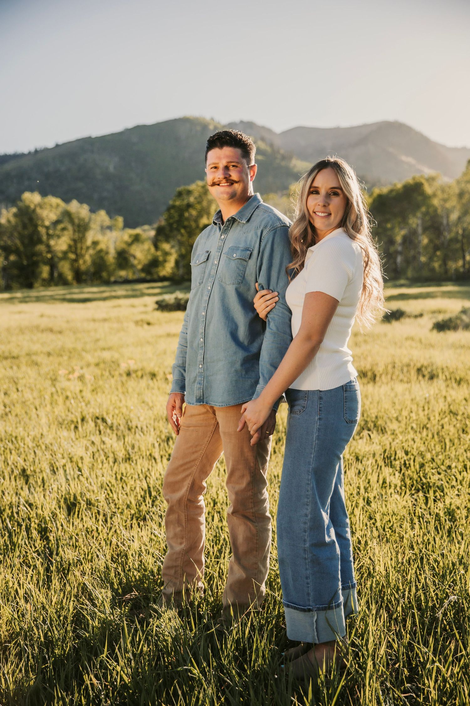 A romantic couple embraces in a sunlit meadow against mountain backdrop during golden hour.