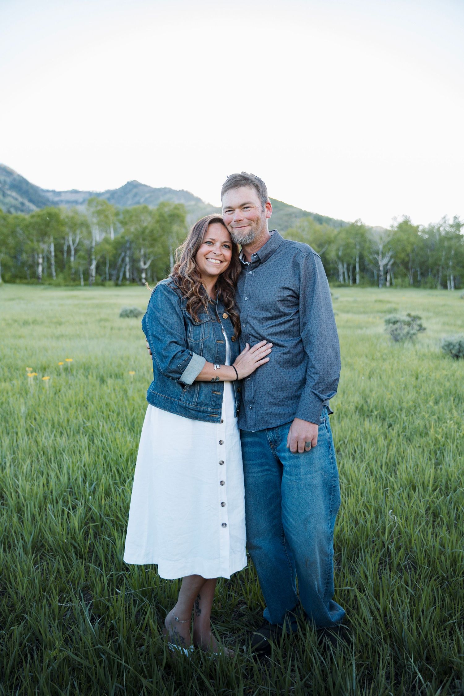 A couple stands close together in a grassy meadow with mountains and aspen trees in the background.